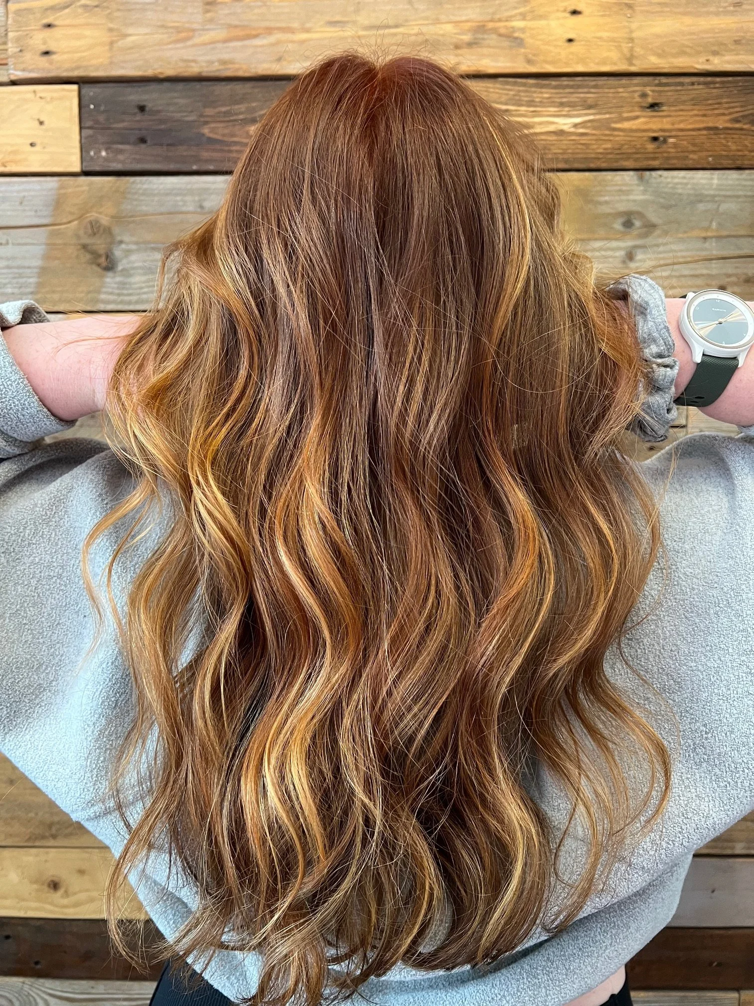 A person with long, wavy red hair seated against a wooden wall.