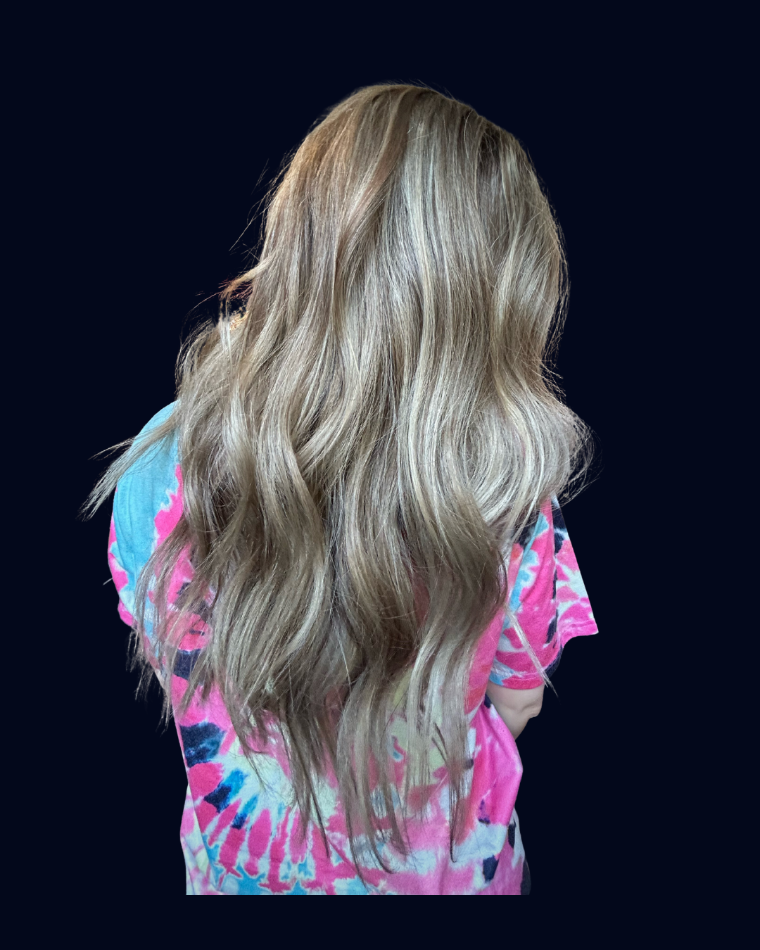 Back view of a woman with long, wavy blonde hair, wearing a colorful, tie-dye pink, blue, and purple t-shirt, against a black background.