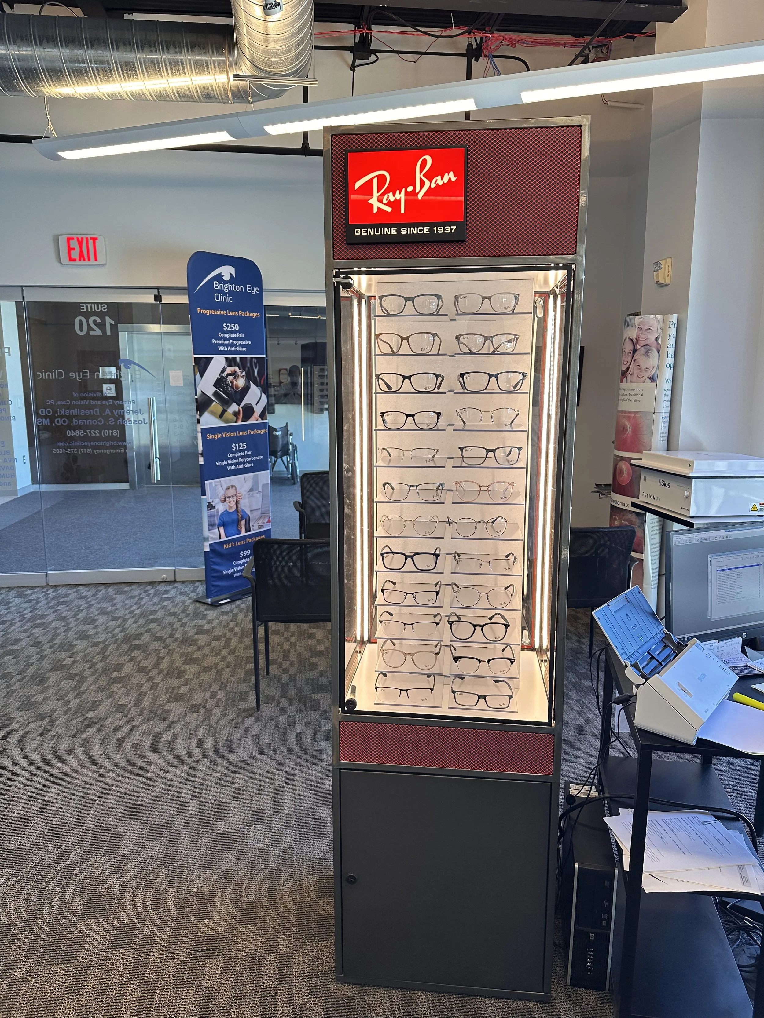 A display case with multiple pairs of eyeglasses inside, located near a desk with a computer and papers, in an office or retail space.