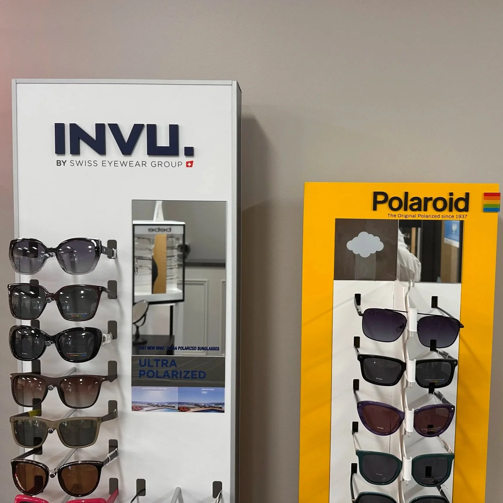 Display of various sunglasses on a stand in an eyewear store, with packaging for INVU and Polaroid sunglasses visible.