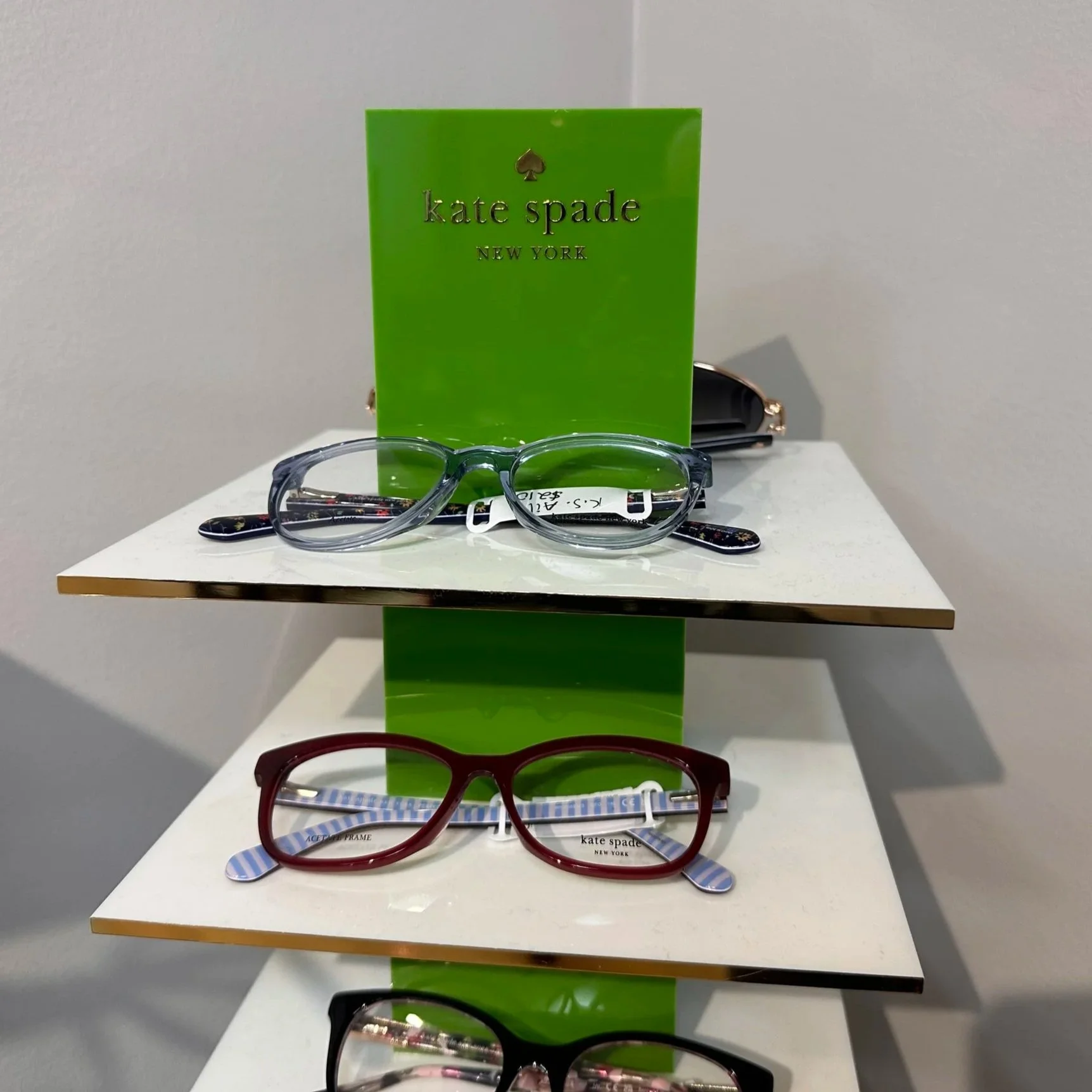 Display of four pairs of Kate Spade eyeglasses on a white shelf, with a green card behind the top pair, and a gray wall in the background.