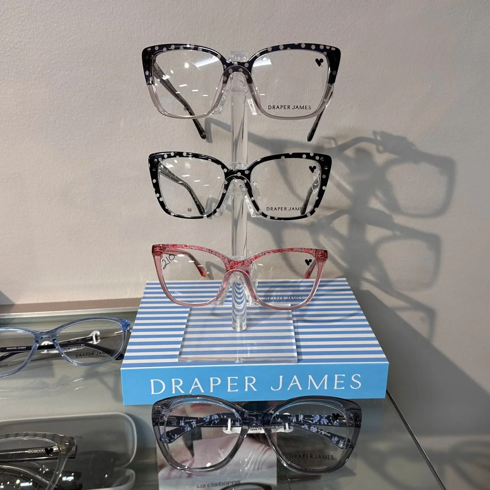 Display of four pairs of designer sunglasses on a stand, with a blue and white striped box labeled 'Draper James' at the base. The sunglasses feature different frame styles and patterns, including black and white polka dots, floral, and clear frames.
