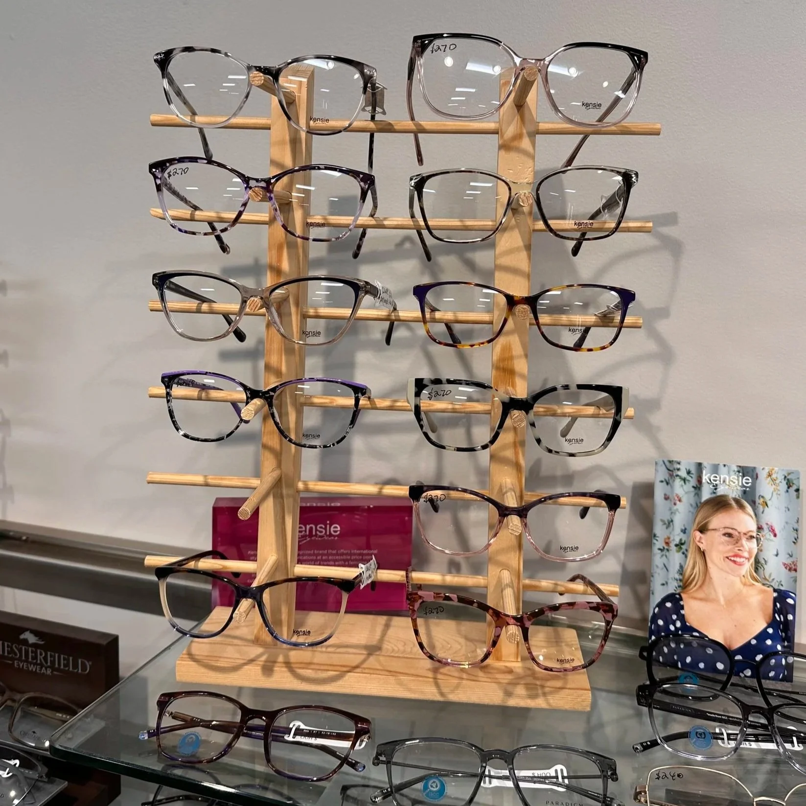 Display of multiple pairs of eyeglasses on a wooden stand at an eyewear store, with a promotional photo of a woman wearing glasses visible in the background.