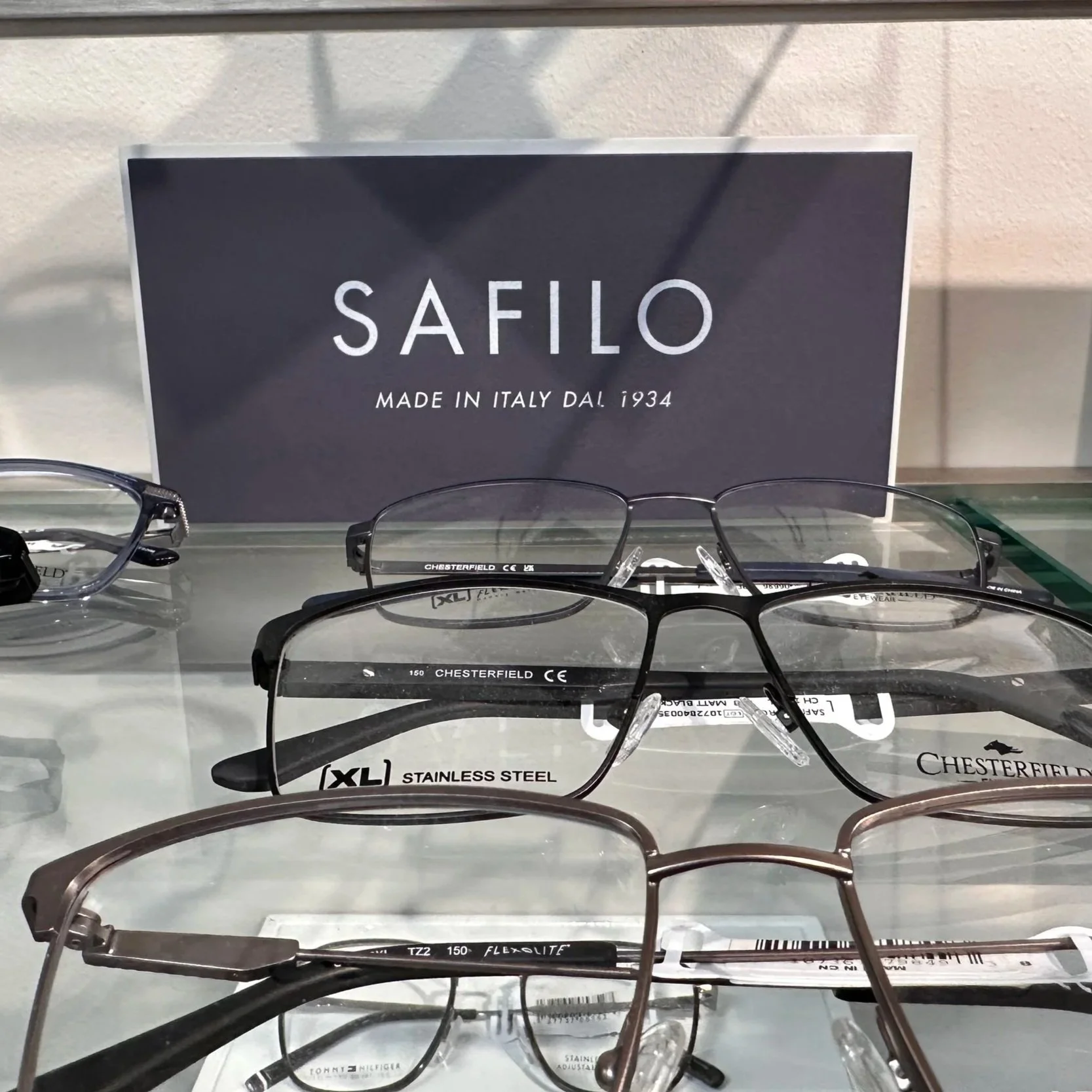 Display of various eyeglasses on a glass shelf in a store, with a sign in the background reading 'Safilo, Made in Italy Daf 1934'.
