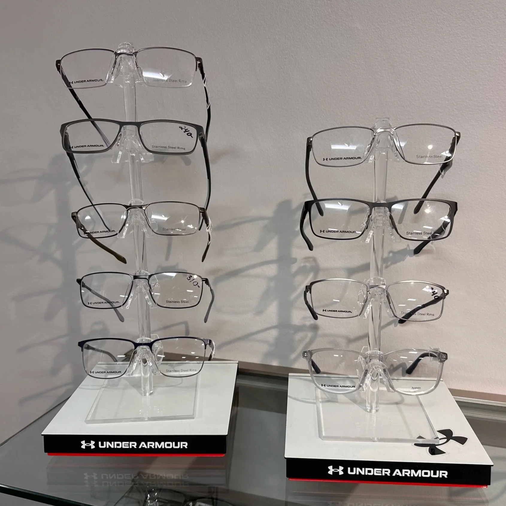 Display of multiple pairs of Under Armour eyeglasses on clear stands in a retail store, arranged in three columns with two rows, on white surfaces with Under Armour branding.