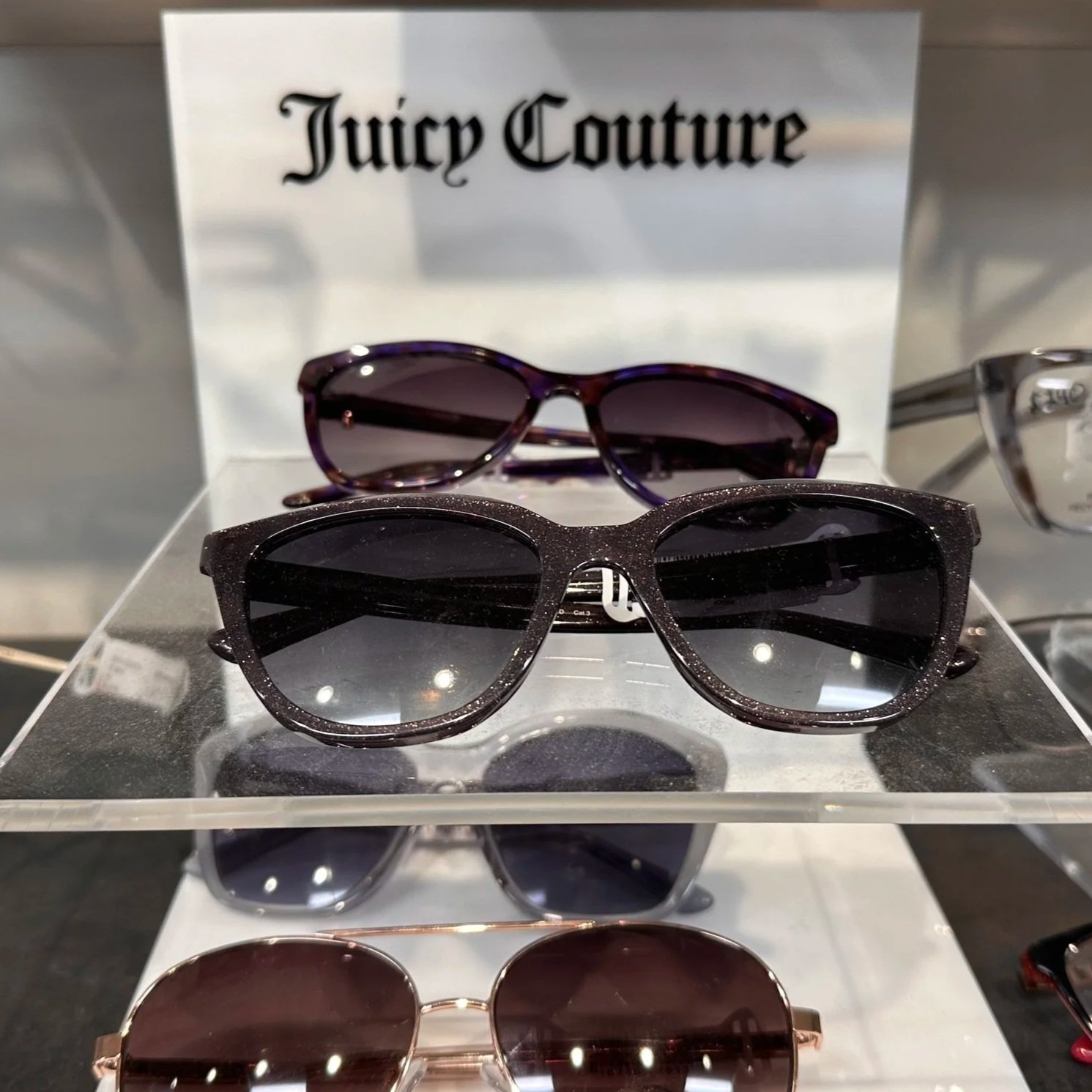 Display of three pairs of Juicy Couture sunglasses on a clear stand with a Juicy Couture sign in the background.
