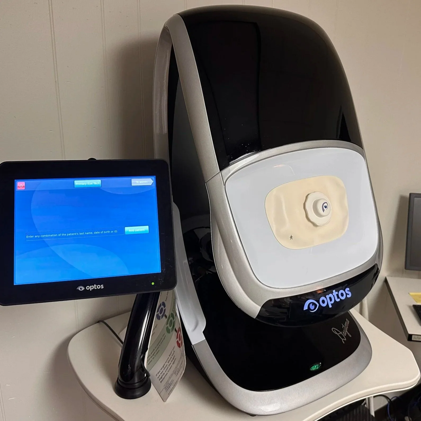 Automated ophthalmology machine used for eye exams, with a monitor displaying patient information and control panel, positioned on a white table in a clinical setting.