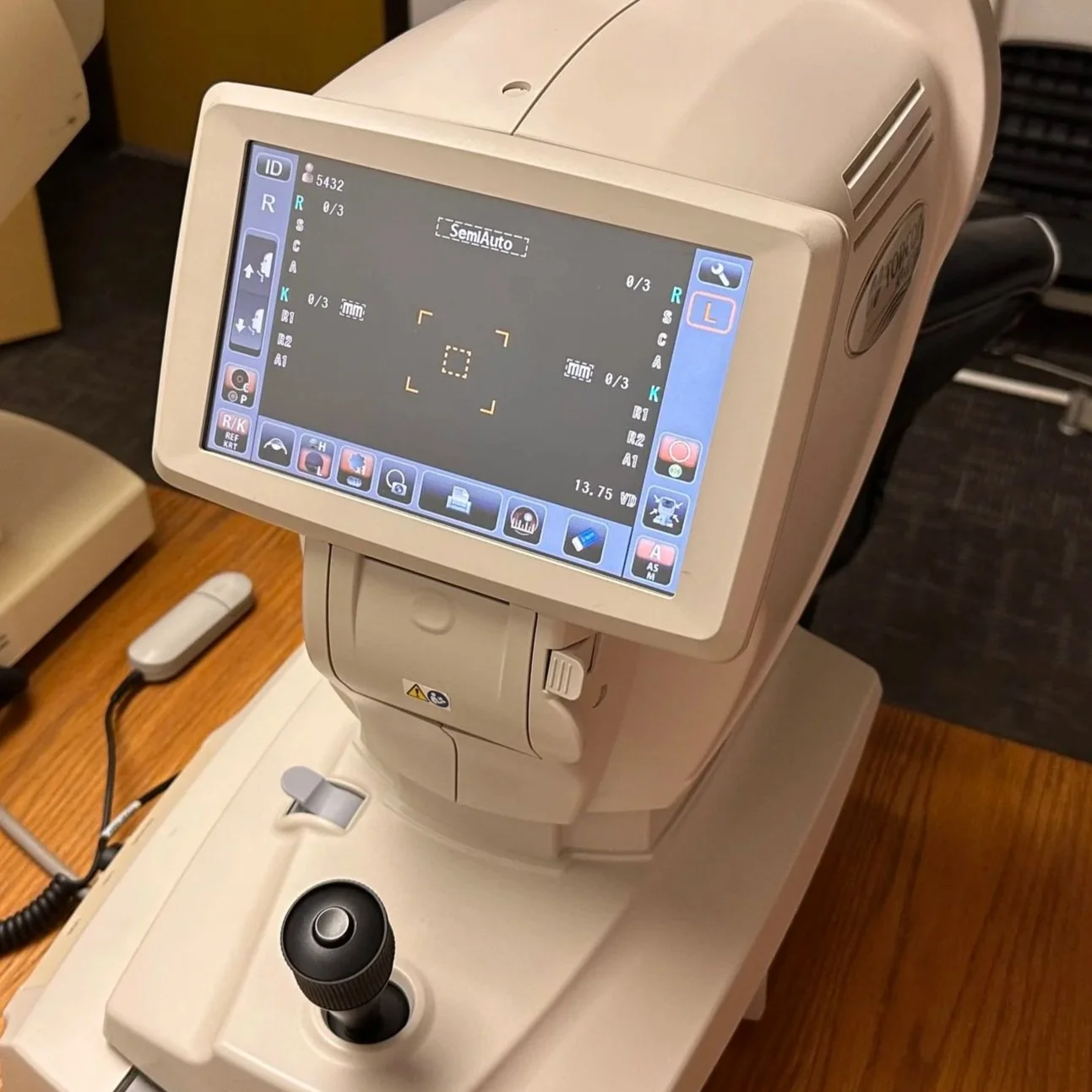 A modern medical machine with a digital display screen, various controls, a joystick, and a foot pedal, likely used for eye examinations or optometry.