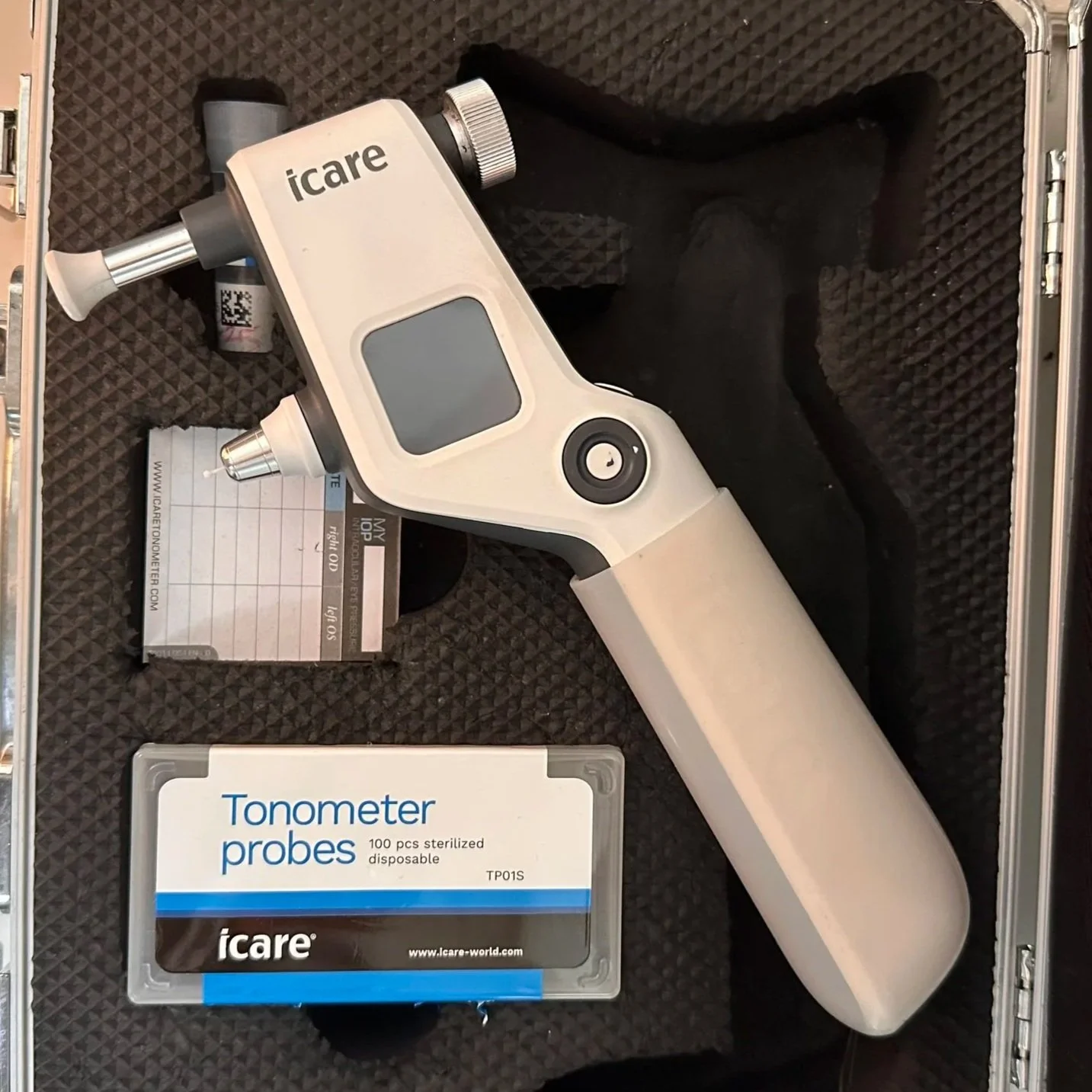 A handheld device used for measuring blood oxygen levels, with a display screen and a probe attachment, placed inside a protective case with a box of disposable tonometer probes nearby.