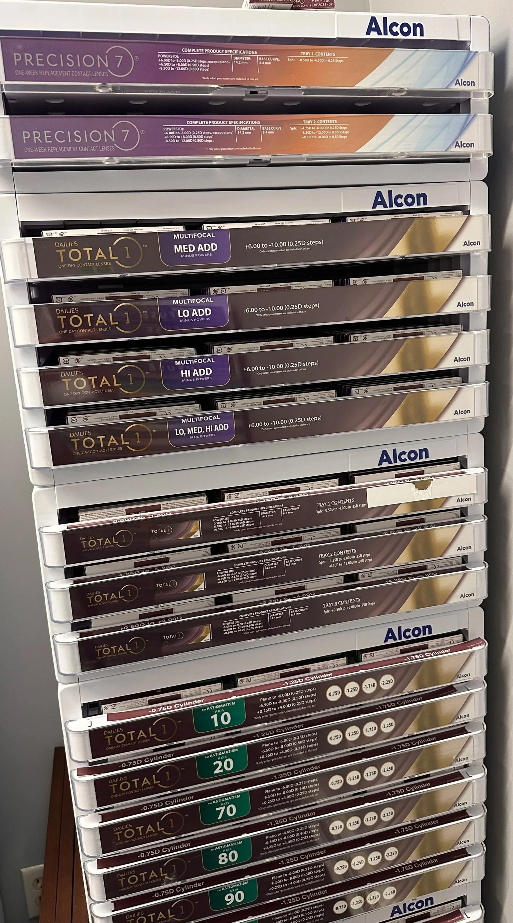 A plastic shelf filled with various boxes of contact lenses from the brand Alcon, including Precision 7, Total 1, and other types of daily contact lenses.