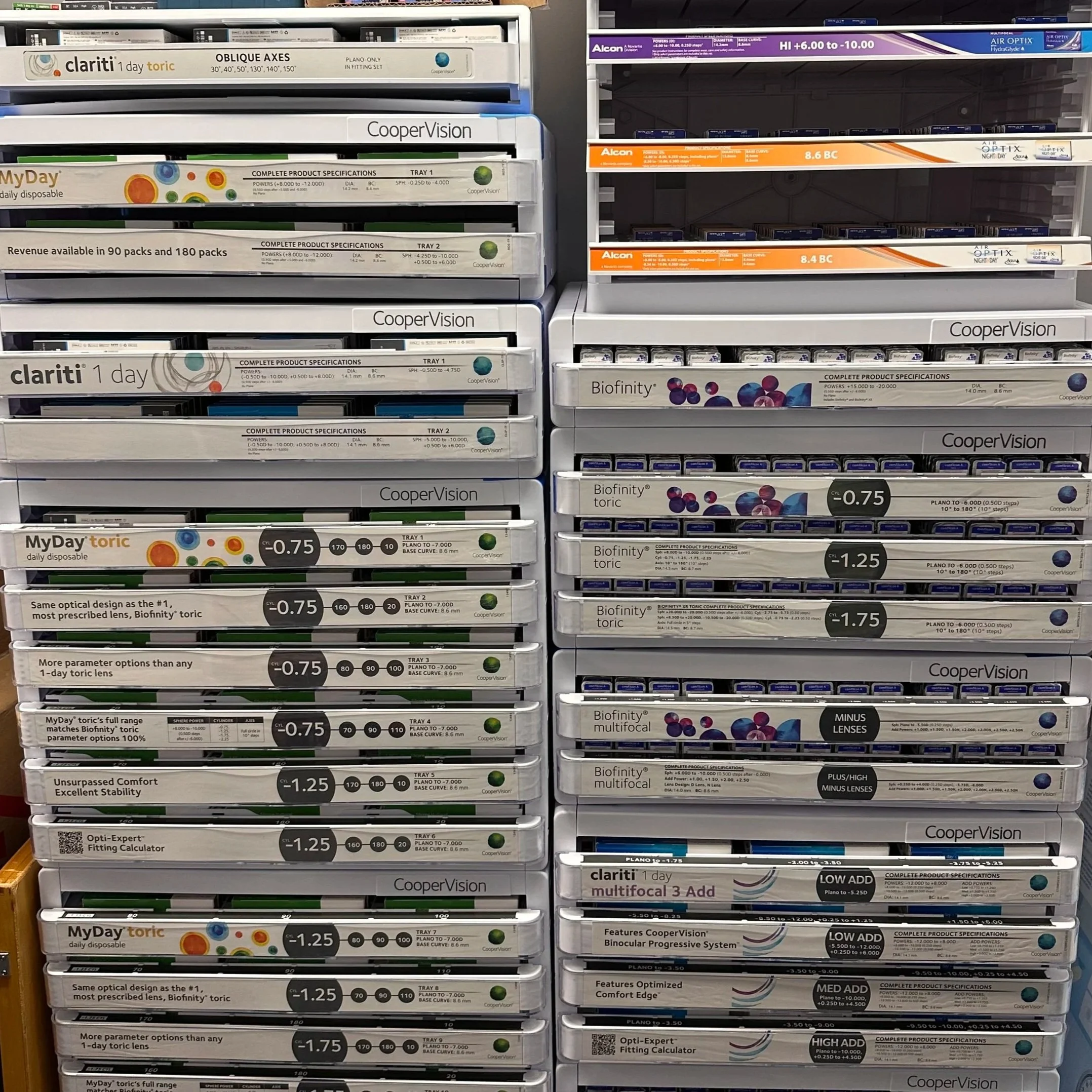 A display of various contact lenses in boxes on store shelves, including brands like Clariti, Biofinity, MyDay, and copervision, with headphones in the background.