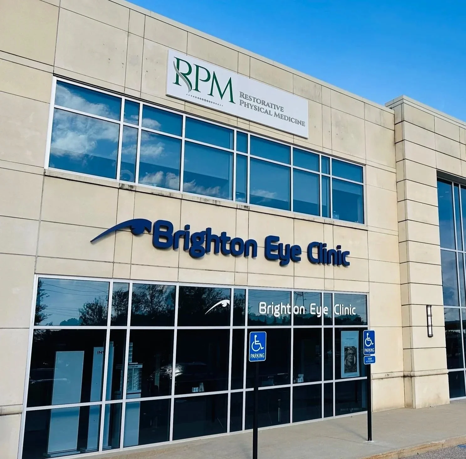 The exterior of a building with signs for Brighton Eye Clinic and RPM Restorative Physical Medicine, blue sky, reflection of clouds in windows, and designated handicapped parking spots in front.
