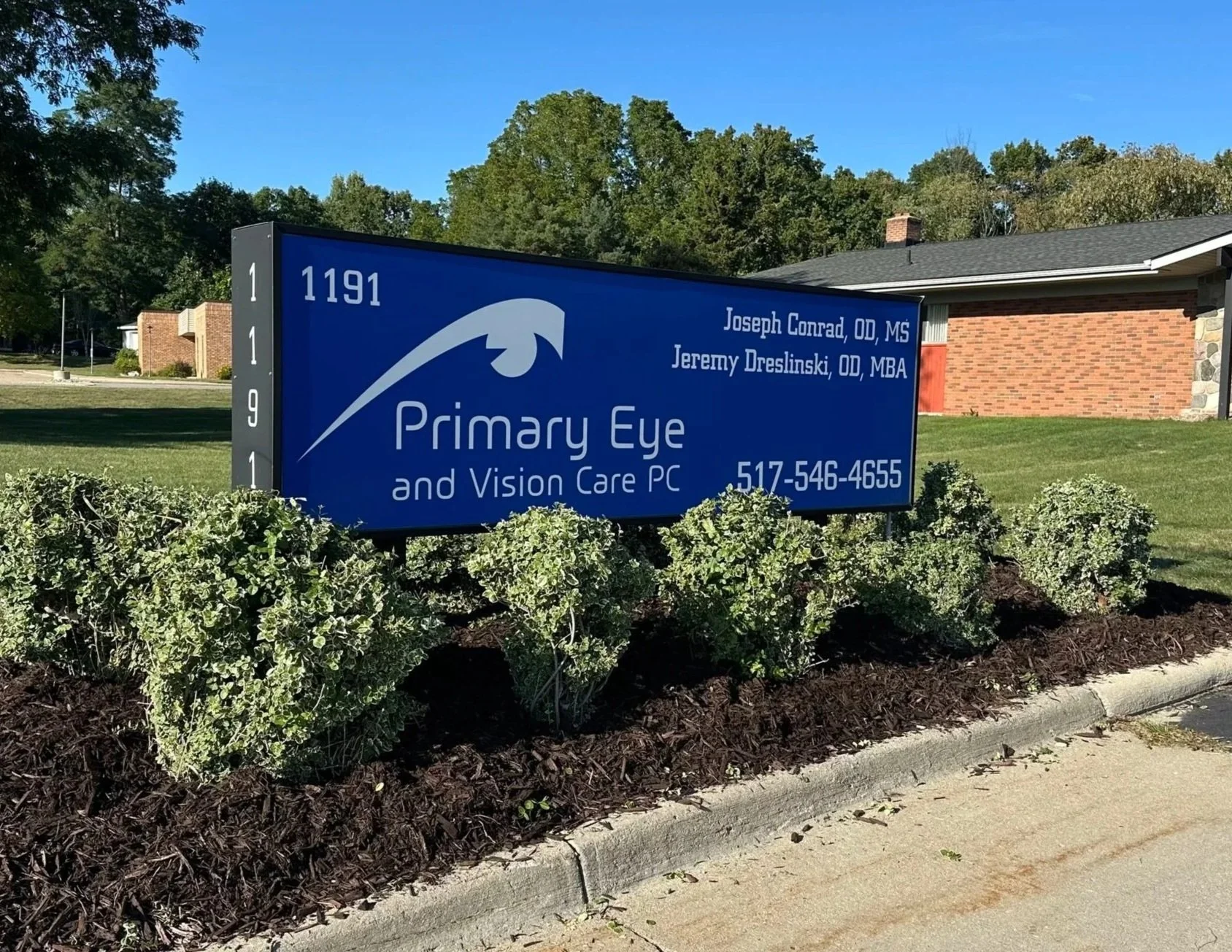 Blue sign for Primary Eye and Vision Care PC with a graphic of an eye, displaying names Joseph Conrad OD MS and Jeremy Dreslinski OD MBA, along with a phone number 517-546-4655, situated in a landscaped area with bushes in front and a brick building 