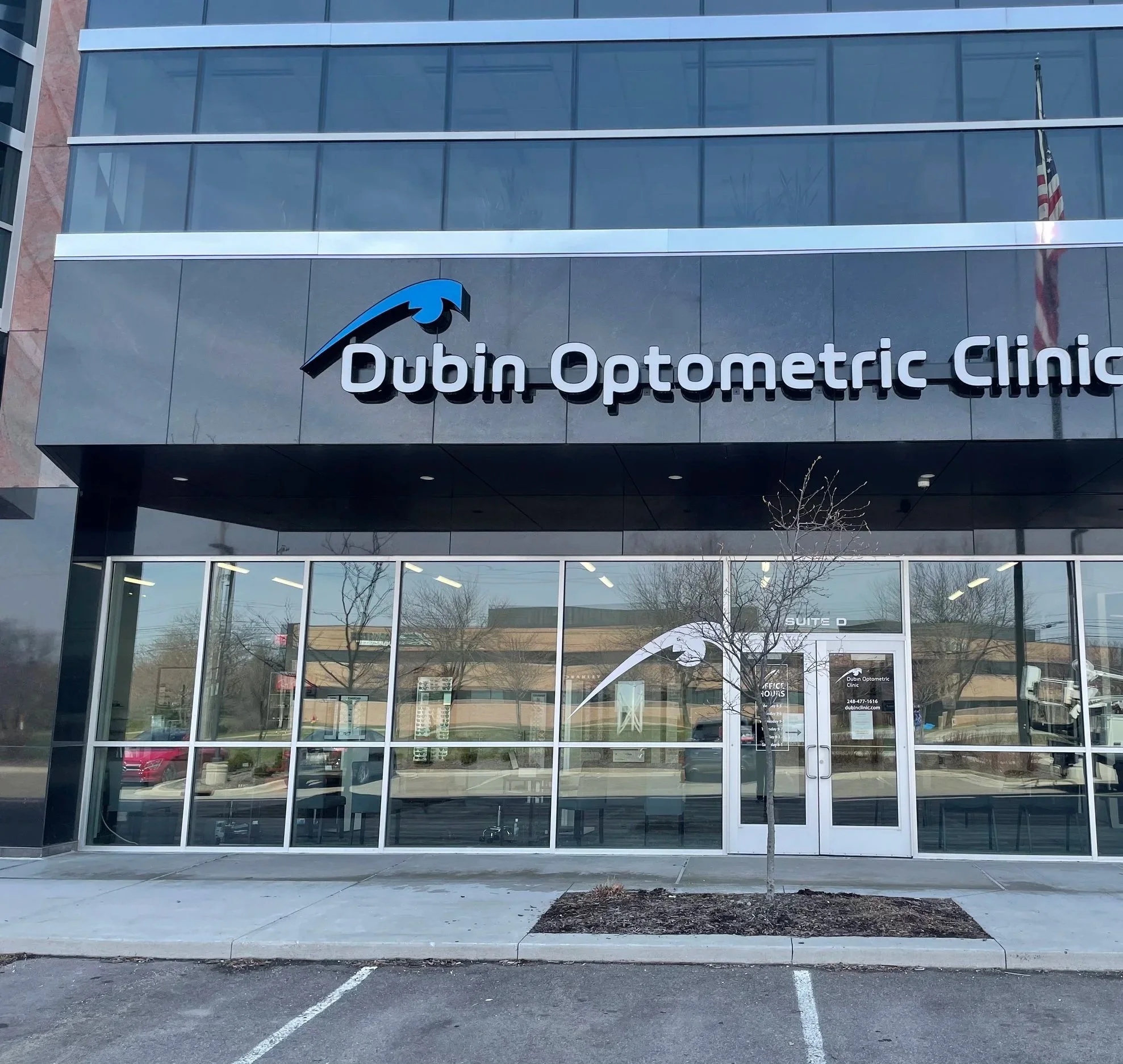 Front view of a modern multi-story building with glass windows and a sign that reads 'Dubin Optometric Clinic' above the entrance. An American flag is visible on the right side of the building.