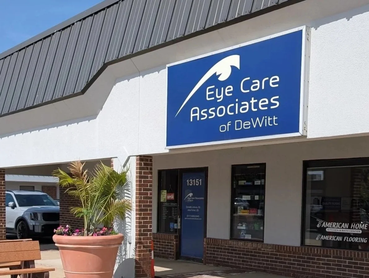 Exterior view of a store named Eye Care Associates of DeWitt with a large blue sign above the entrance. The store has brick and white walls, large windows, and a potted plant outside.