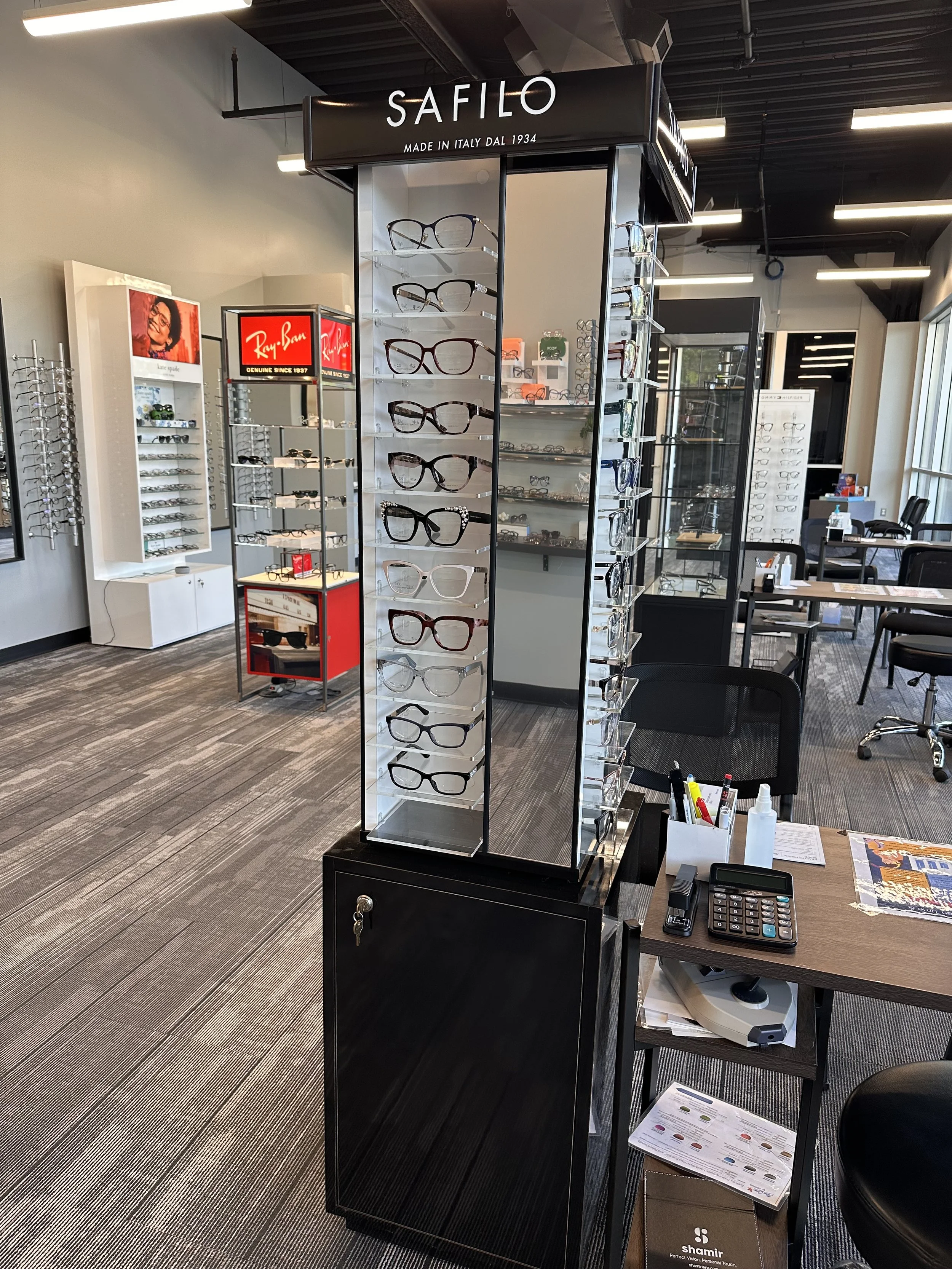 An eyeglasses display stand in an eyewear store with the brand name 'Safilo' and text 'Made in Italy dal 1934' at the top. Surrounding the stand are tables, chairs, and other displays with eyeglasses and accessories.