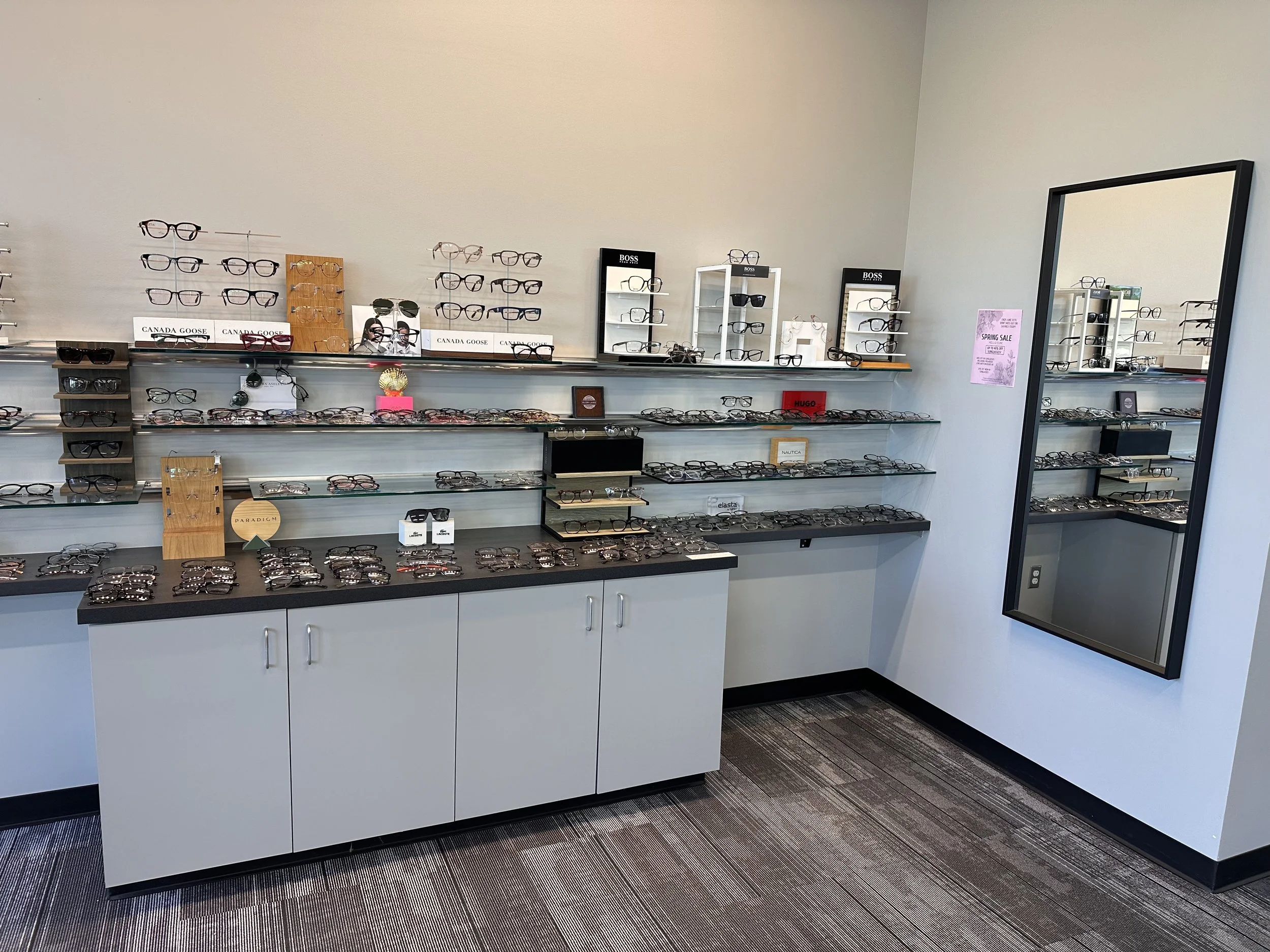 Display of eyeglasses and sunglasses on glass shelves in an eyewear store, with branded boxes and signage, and a large mirror on the right side.