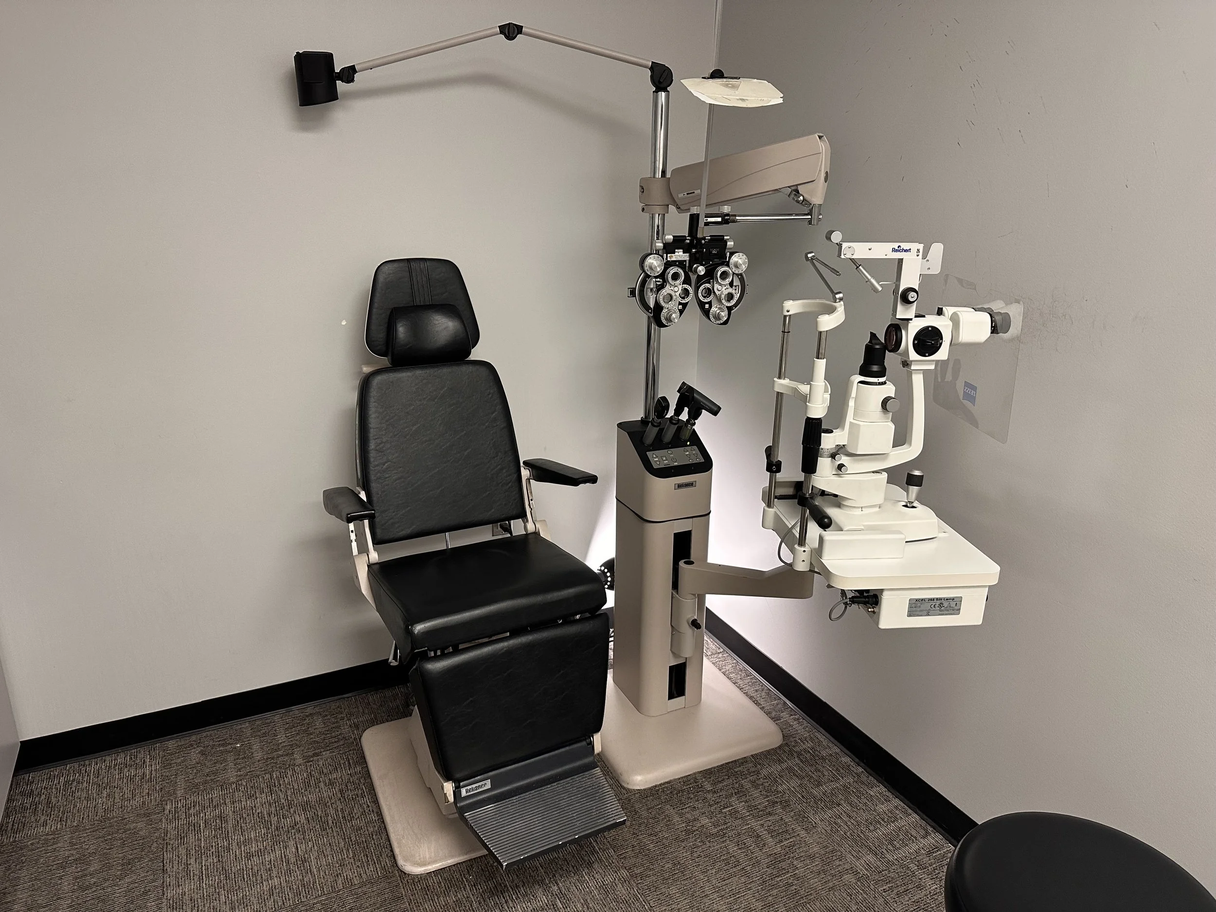 An eye examination room with a black ophthalmic examination chair, specialized eye examination equipment, and a plain wall.