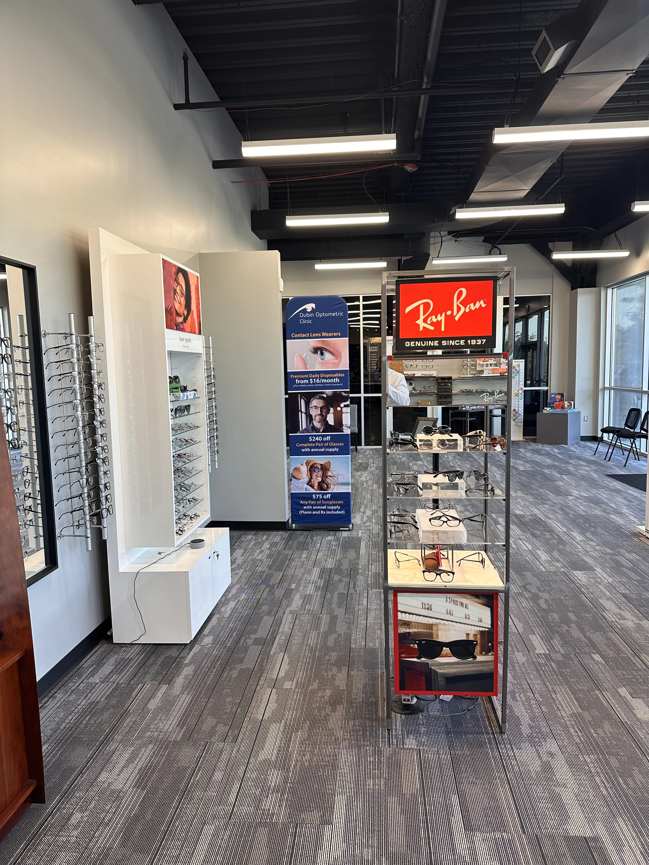 Inside an eyewear store with displays of glasses on lines on the wall and a free-standing rack with sunglasses. A Ray-Ban sign and promotional banner are visible.