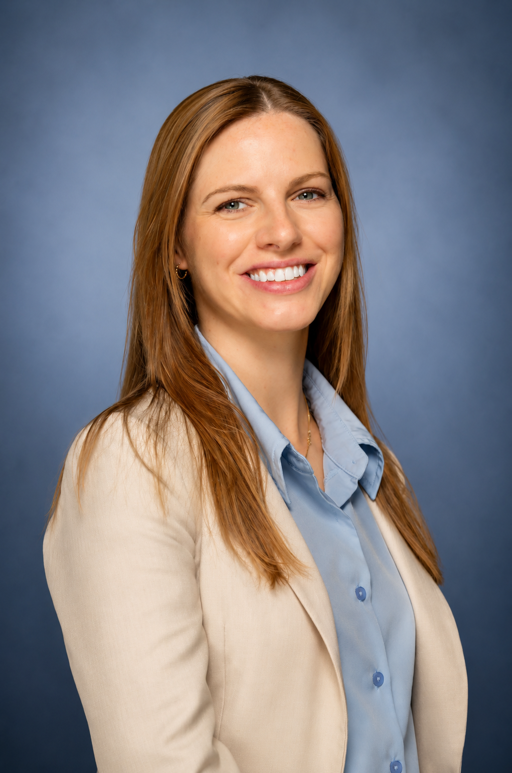 A woman with long red hair wearing a beige blazer and light blue shirt, smiling in front of a blue background.