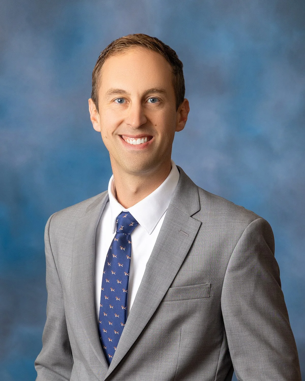 A professional headshot of a young man with short brown hair, wearing a gray suit, white shirt, and a navy tie with small white patterns, smiling against a blue background.