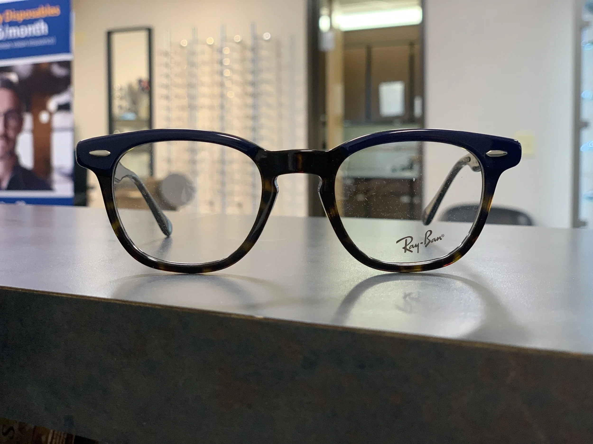 A pair of Ray-Ban eyeglasses resting on a white surface with a blurred background of what appears to be an optometry office or store.