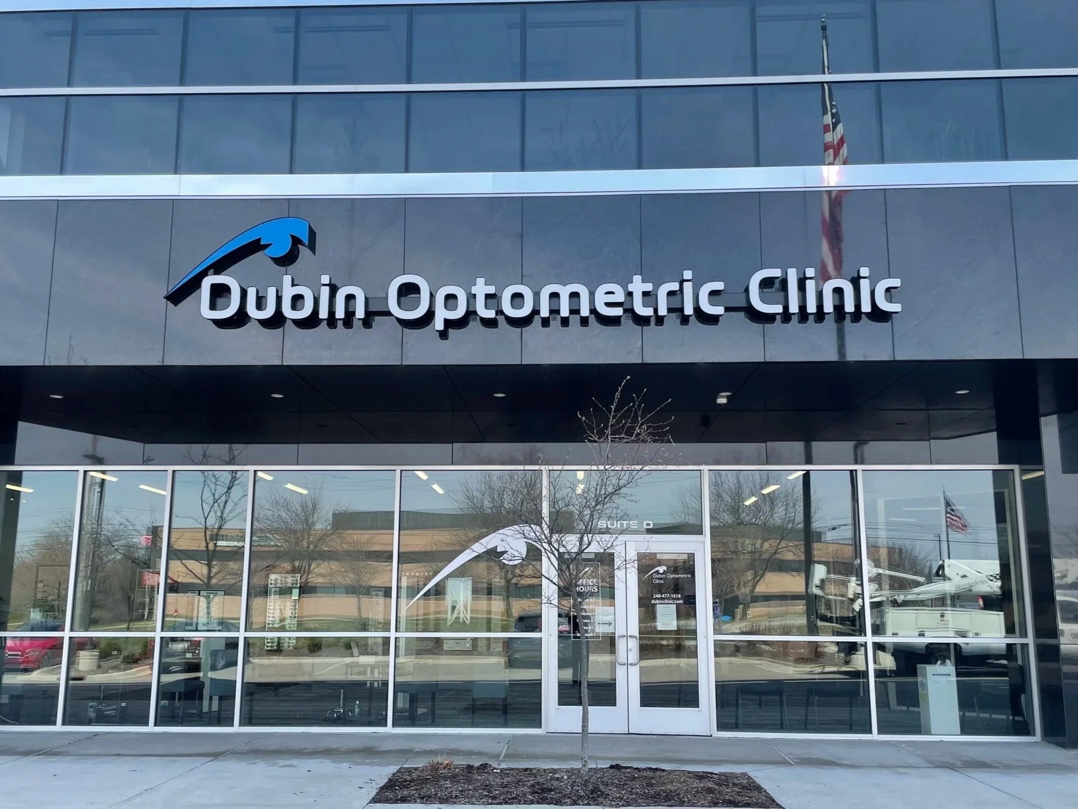 Front view of the Dubin Optometric Clinic building with glass windows, a sign displaying its name and logo, and an American flag reflected in the windows.