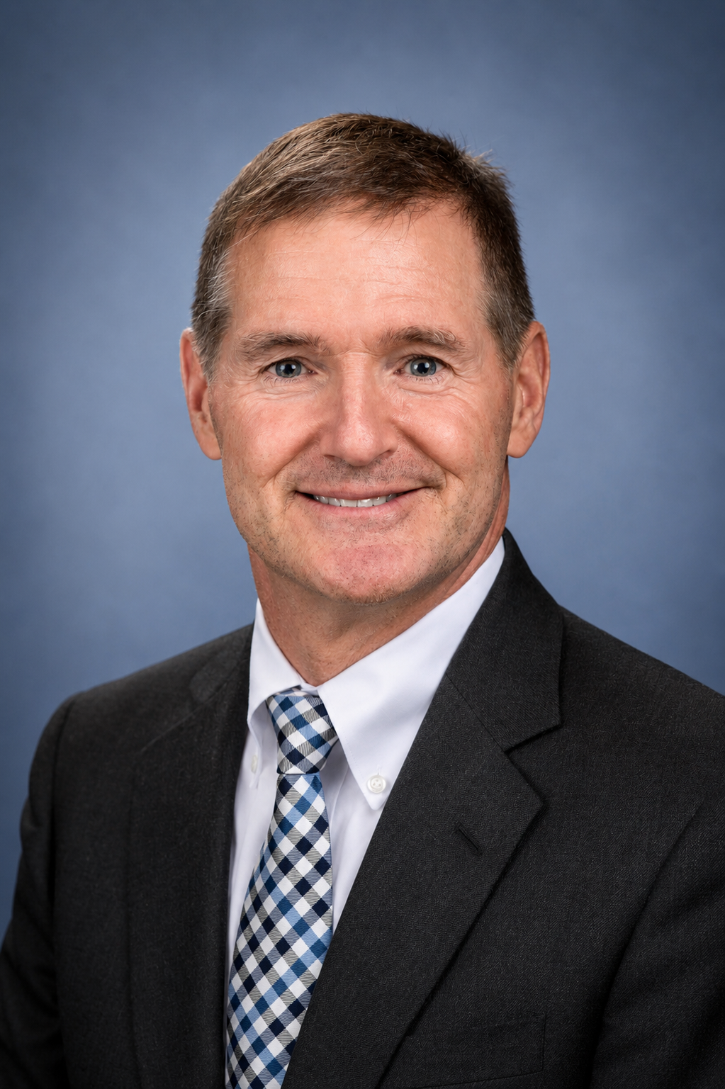 Professional headshot of a smiling middle-aged man with short brown hair, wearing a black suit, white shirt, and patterned blue and white tie, against a blue gradient background.