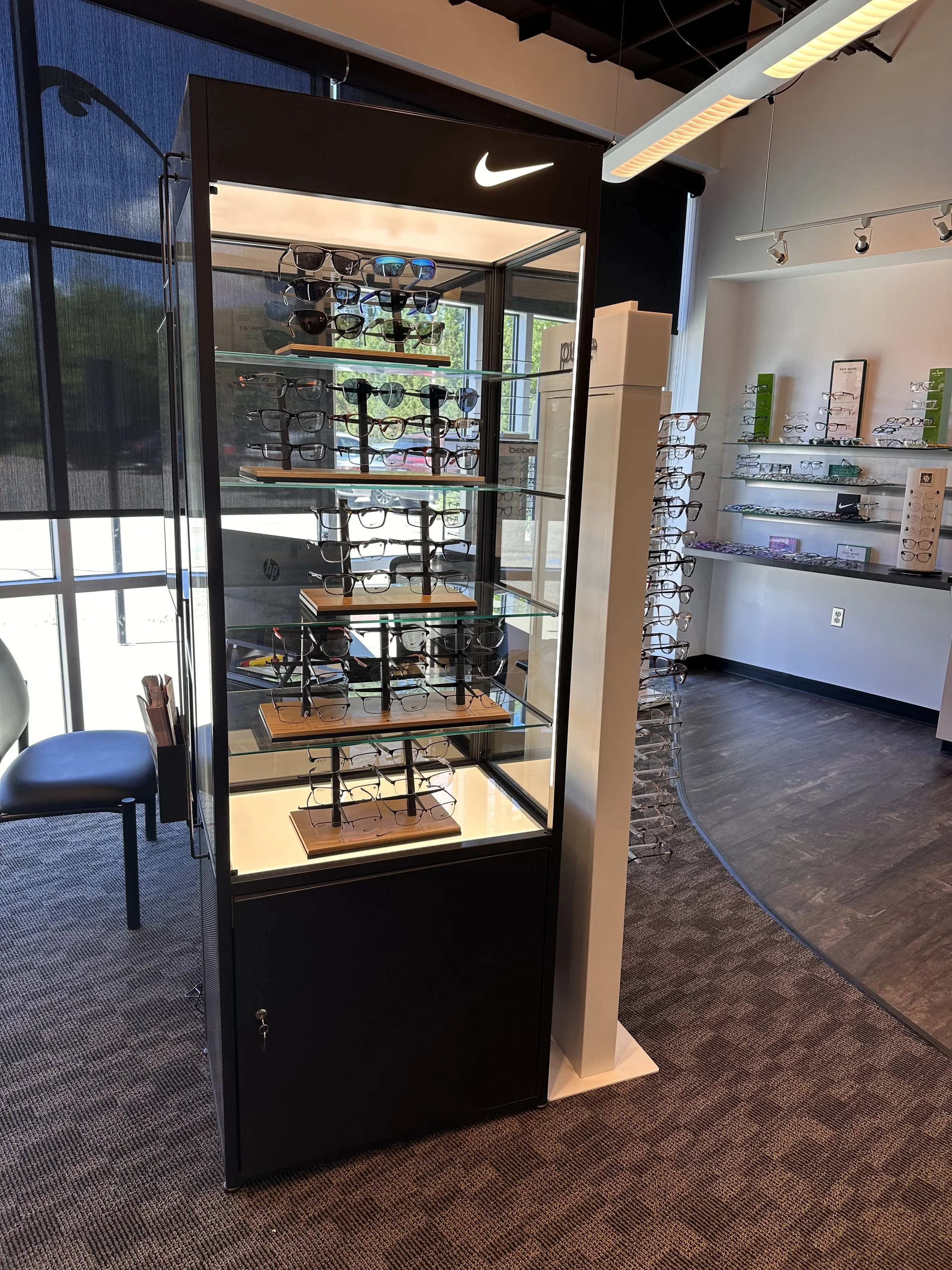 A display case with multiple shelves holding sunglasses inside a store, with a large Nike logo at the top, and other eyewear displays on the wall in the background.