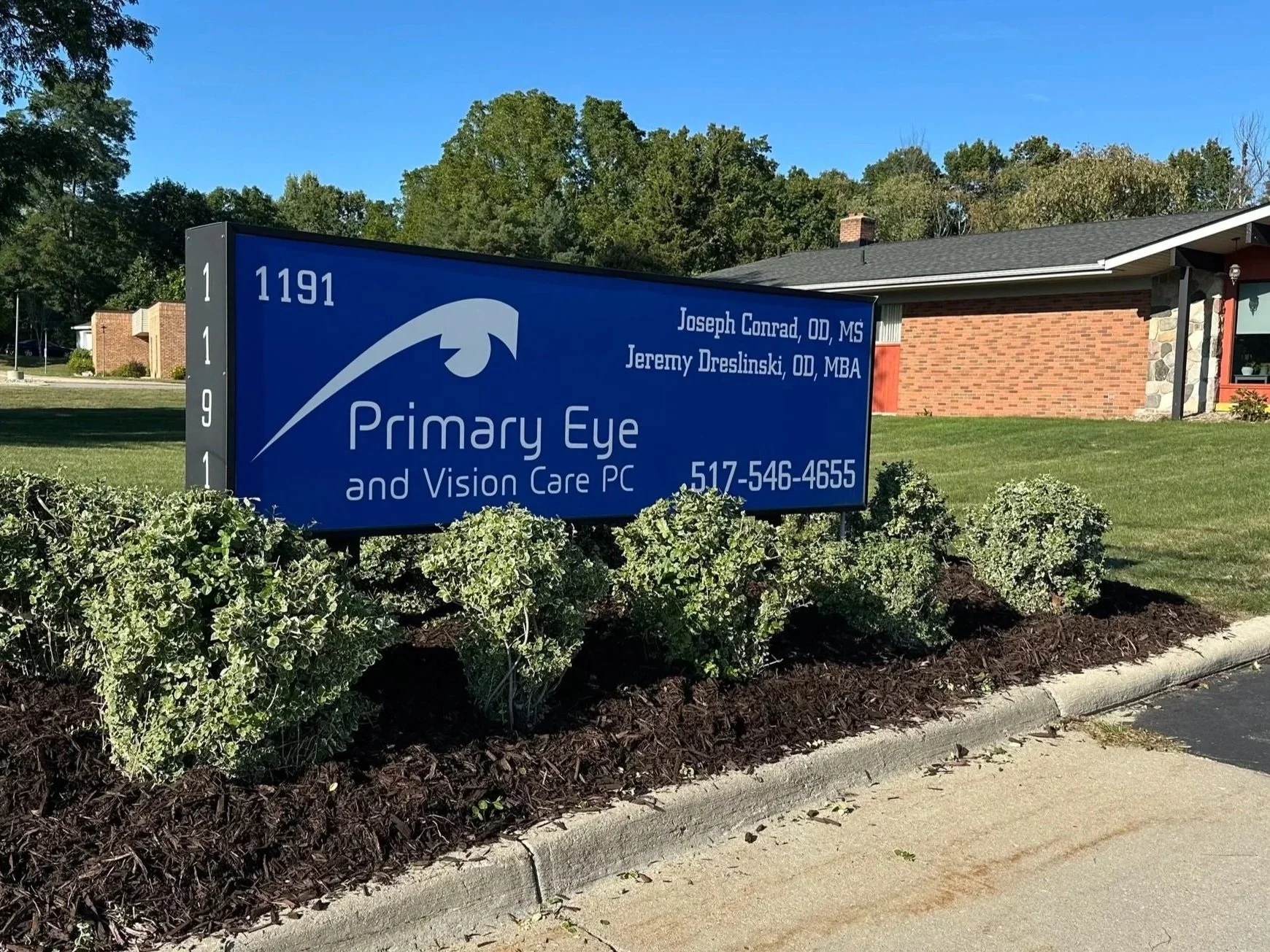 Blue sign for Primary Eye and Vision Care PC with name and contact information, placed in front of a brick building with a lawn and bushes, under a clear blue sky.