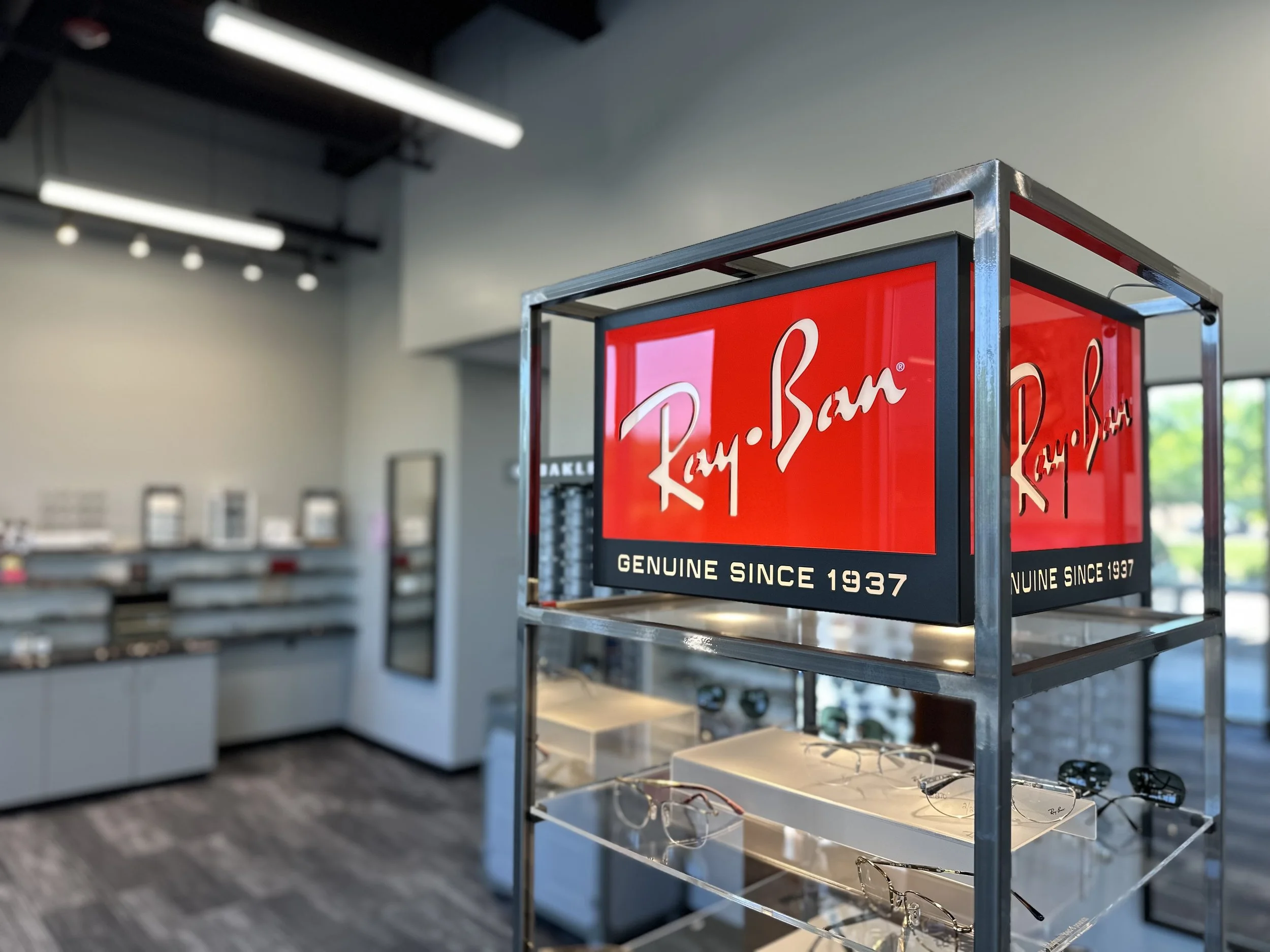 A retail display stand with sunglasses and eyeglasses next to a red sign reading "Ray-Ban," indicating the brand, with additional text "Genuine Since 1937," inside an eyewear store.