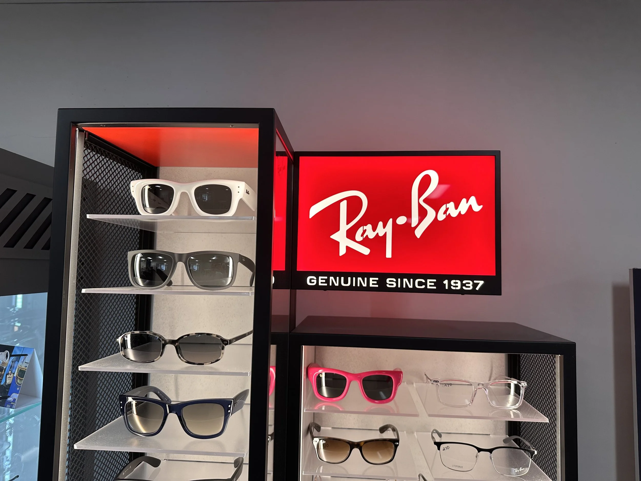 Display of Ray-Ban sunglasses on shelves, with a red Ray-Ban sign and slogan 'Genuine Since 1937' in the background.