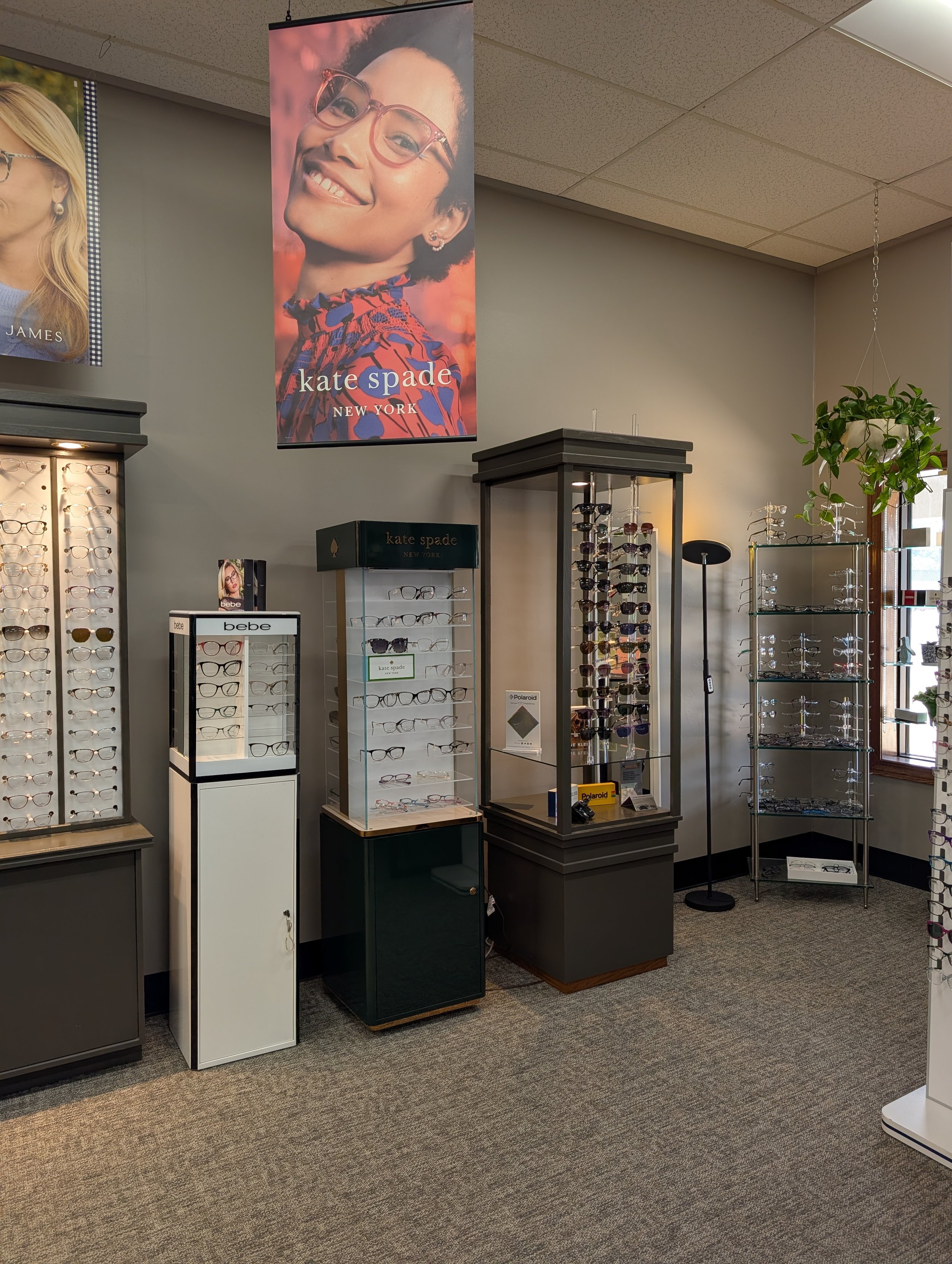 Interior of an eyewear store displaying various eyeglasses on glass and wooden display racks, with a large banner of a woman wearing glasses from Kate Spade New York hanging on the wall.