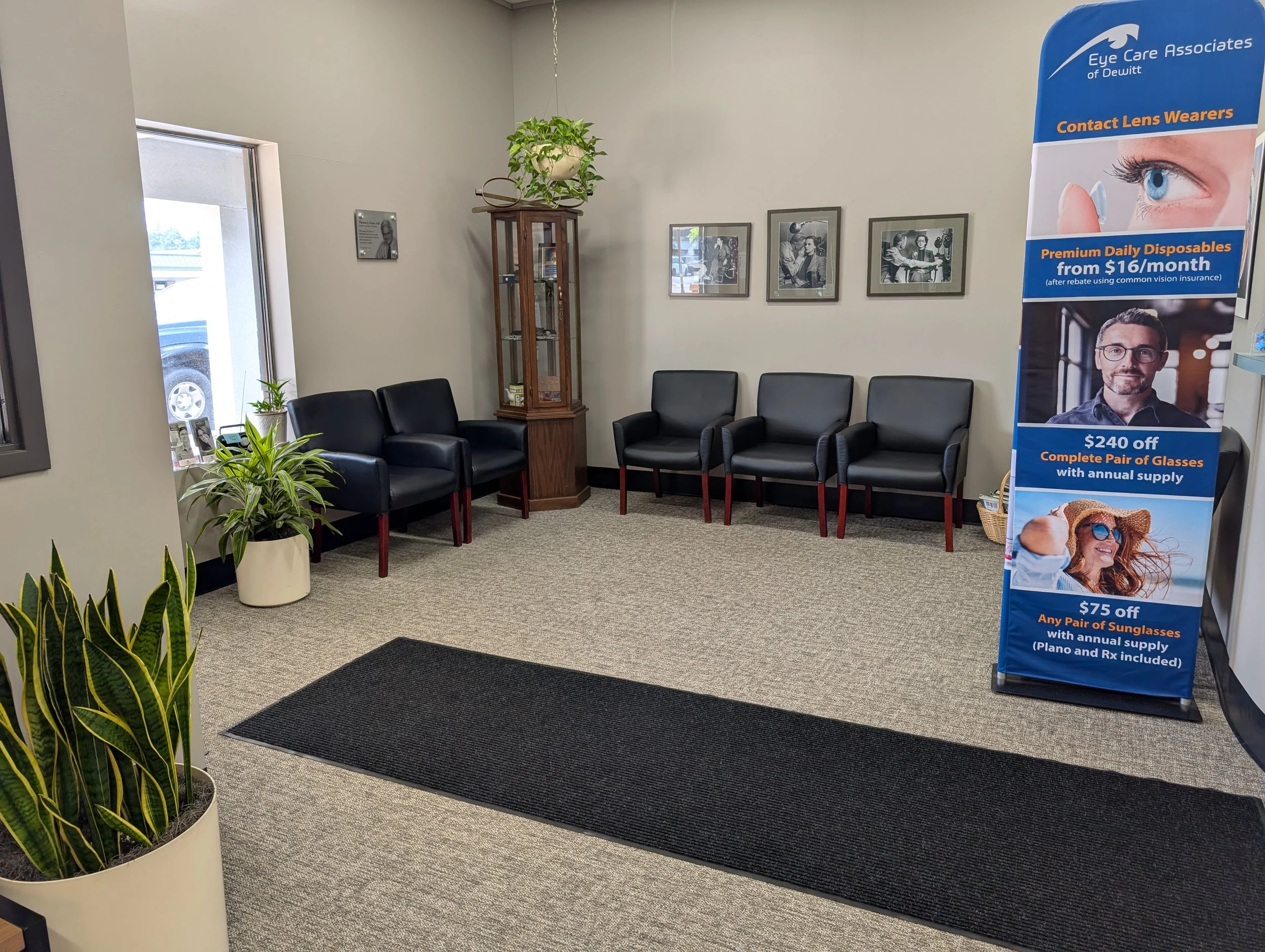 An waiting area inside an eye care clinic with black chairs, potted plants, and framed photographs on the wall. There is a tall wooden display case with a hanging plant on top, and a large blue promotional banner advertising glasses and contact lens 