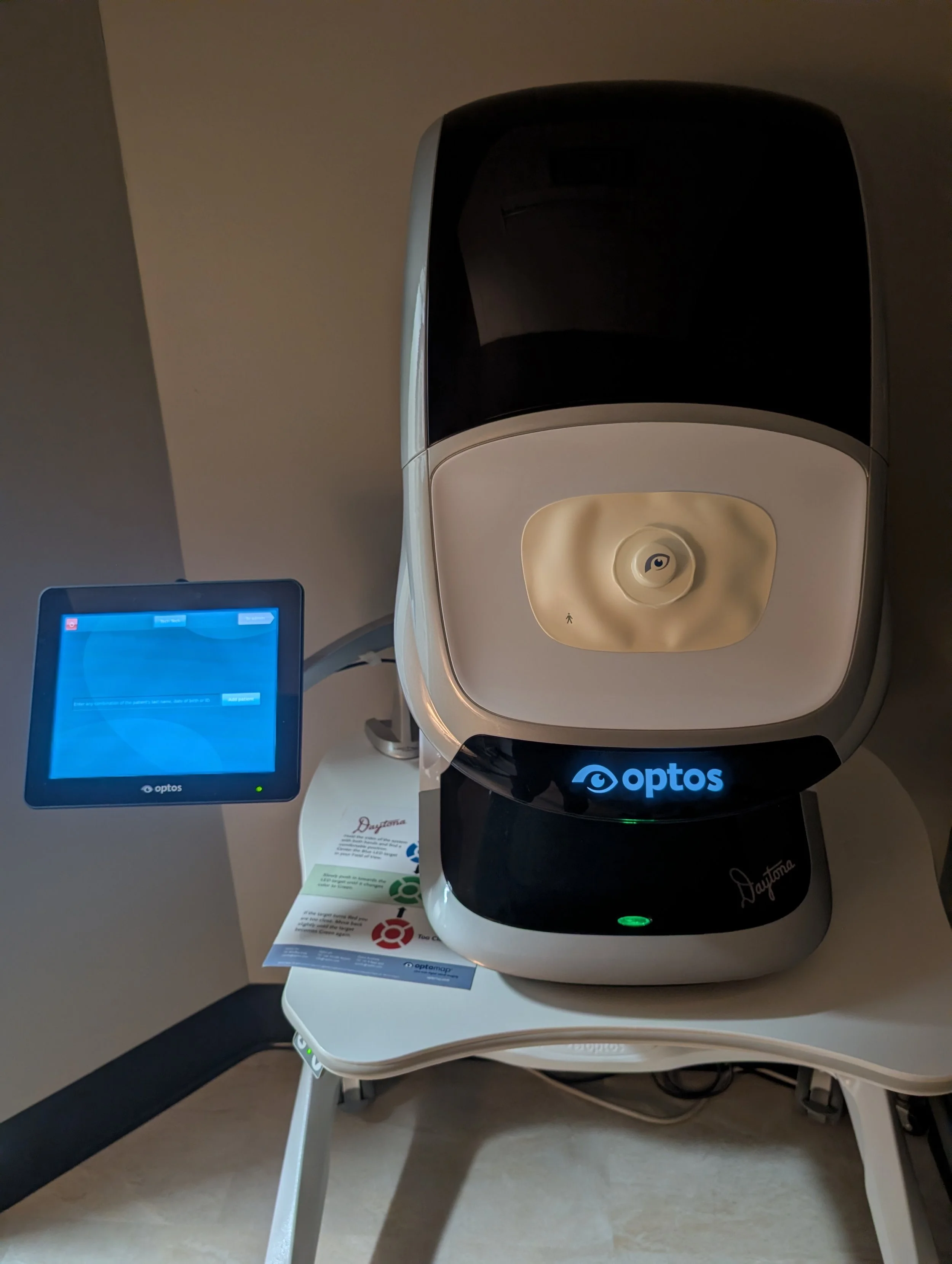 An Optos medical imaging device on a white stand, next to a computer monitor, in a medical clinic setting.