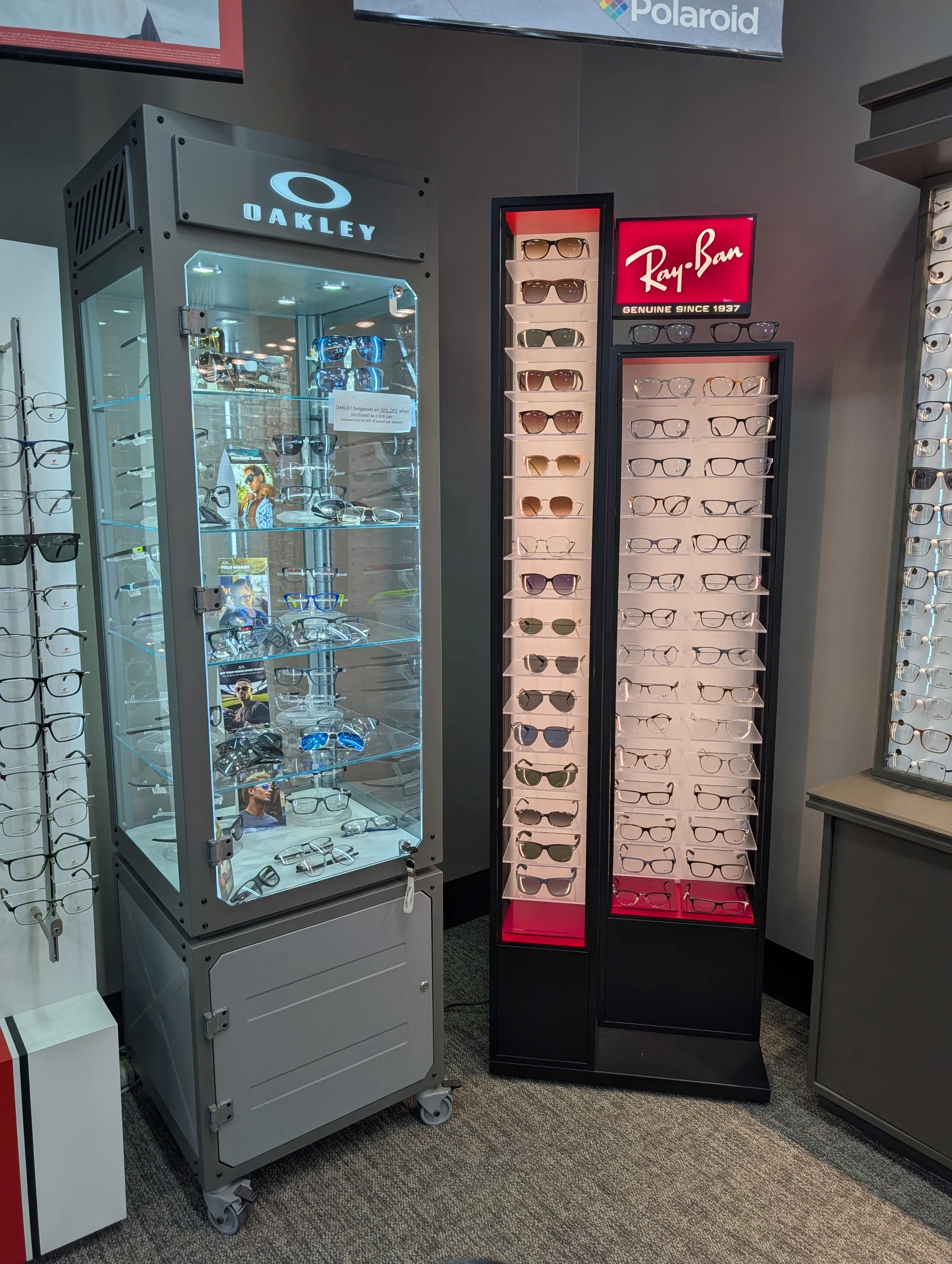 Display of sunglasses and eyeglasses in a store, including a glass cabinet with various eyewear and a tall black stand holding multiple pairs of sunglasses and eyeglasses.