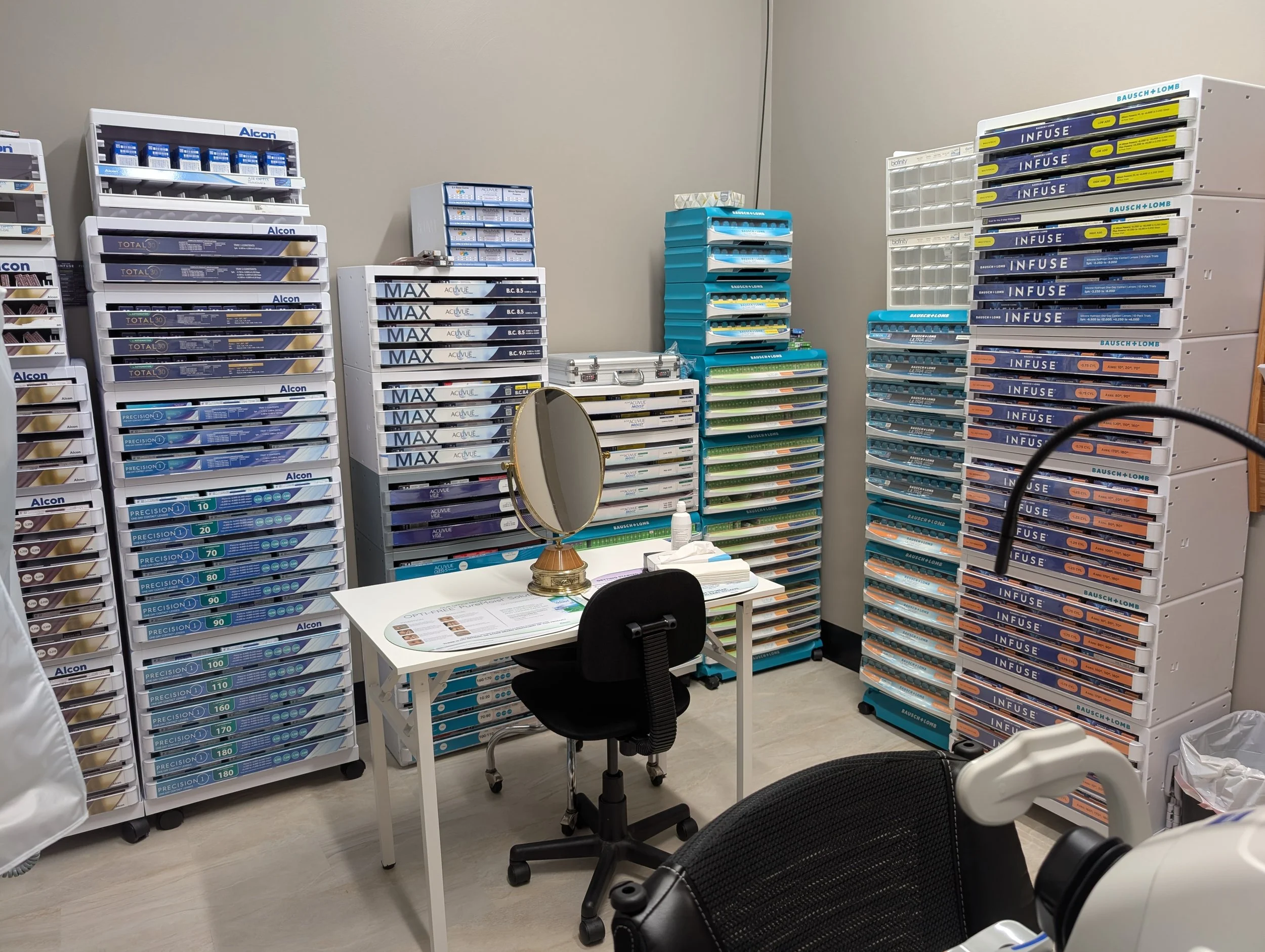 A storage room with shelves filled with various boxes of contact lenses and related products, a small table with a round mirror, and office chairs.