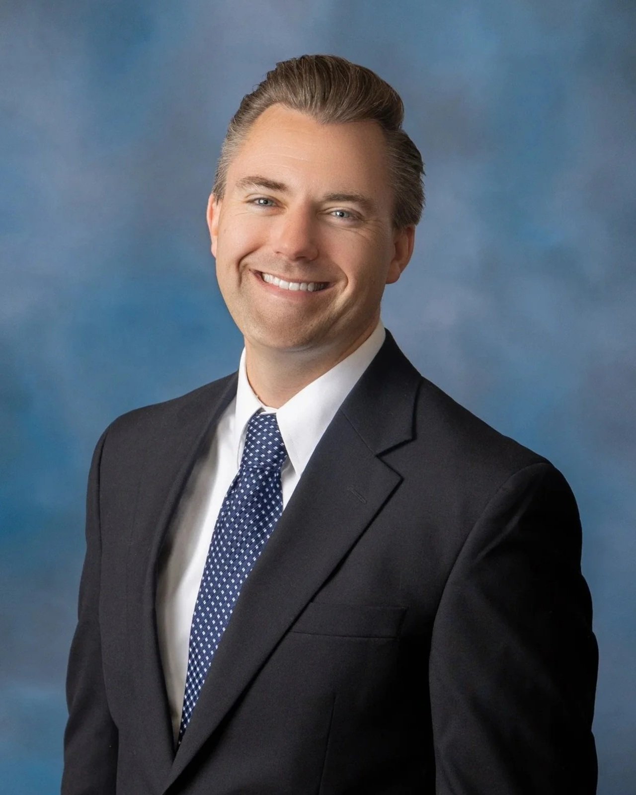 A professional portrait of a man in a suit, smiling, with a blue background.