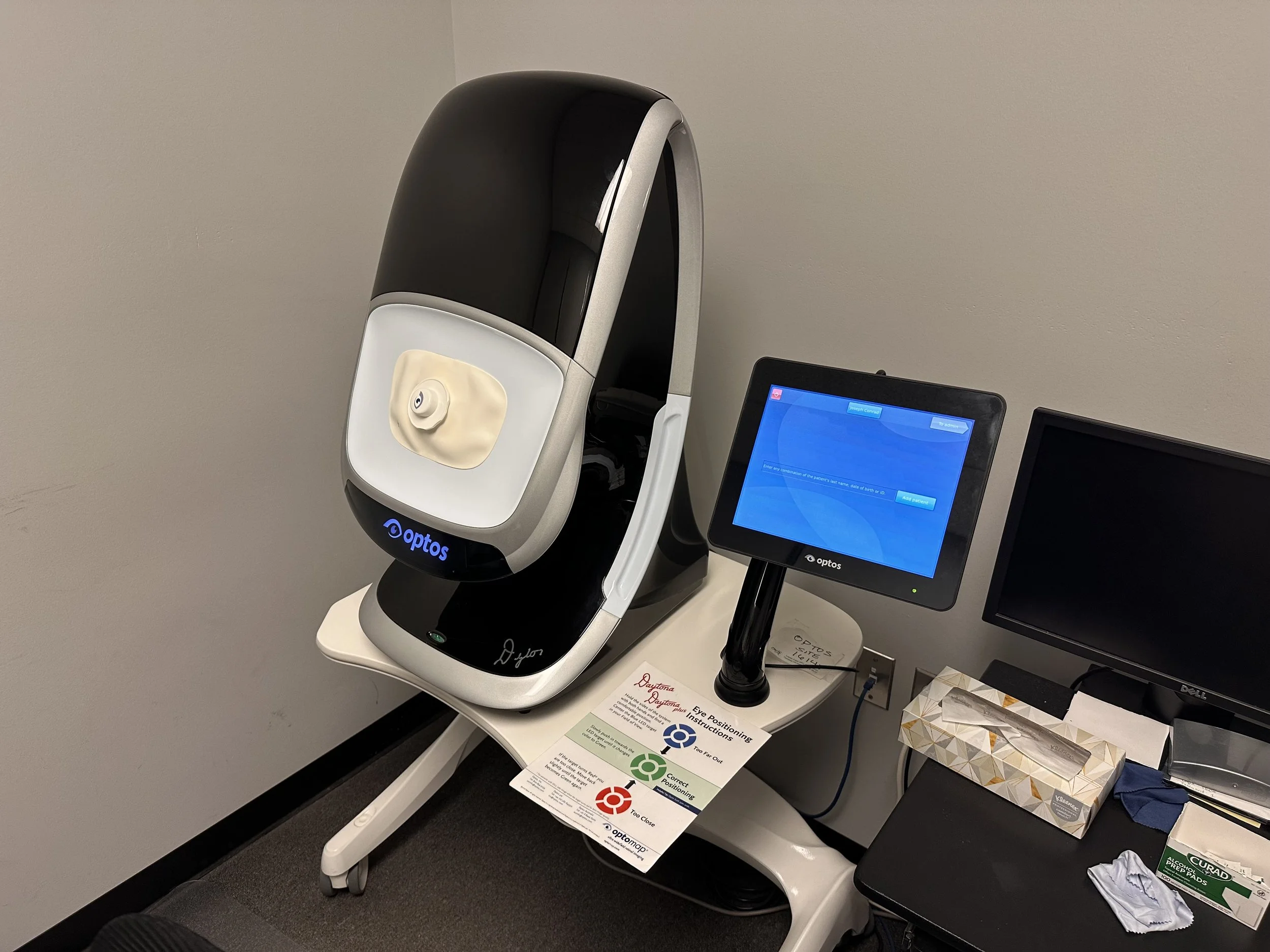Medical equipment on a table in a clinic or hospital, including an optometry device and additional monitor screens, with a sign explaining eye positioning instructions.