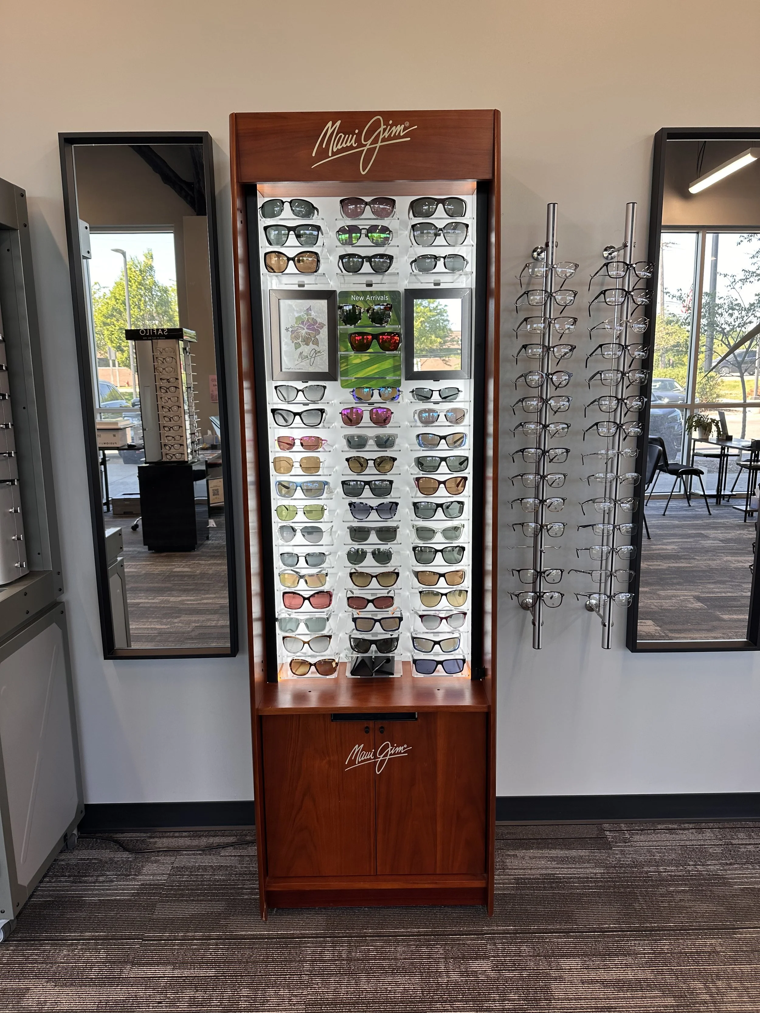 A display case of Maui Jim sunglasses with a wooden frame and a sign that says 'Maui Jim' at the top, located between two large mirrors inside a store.