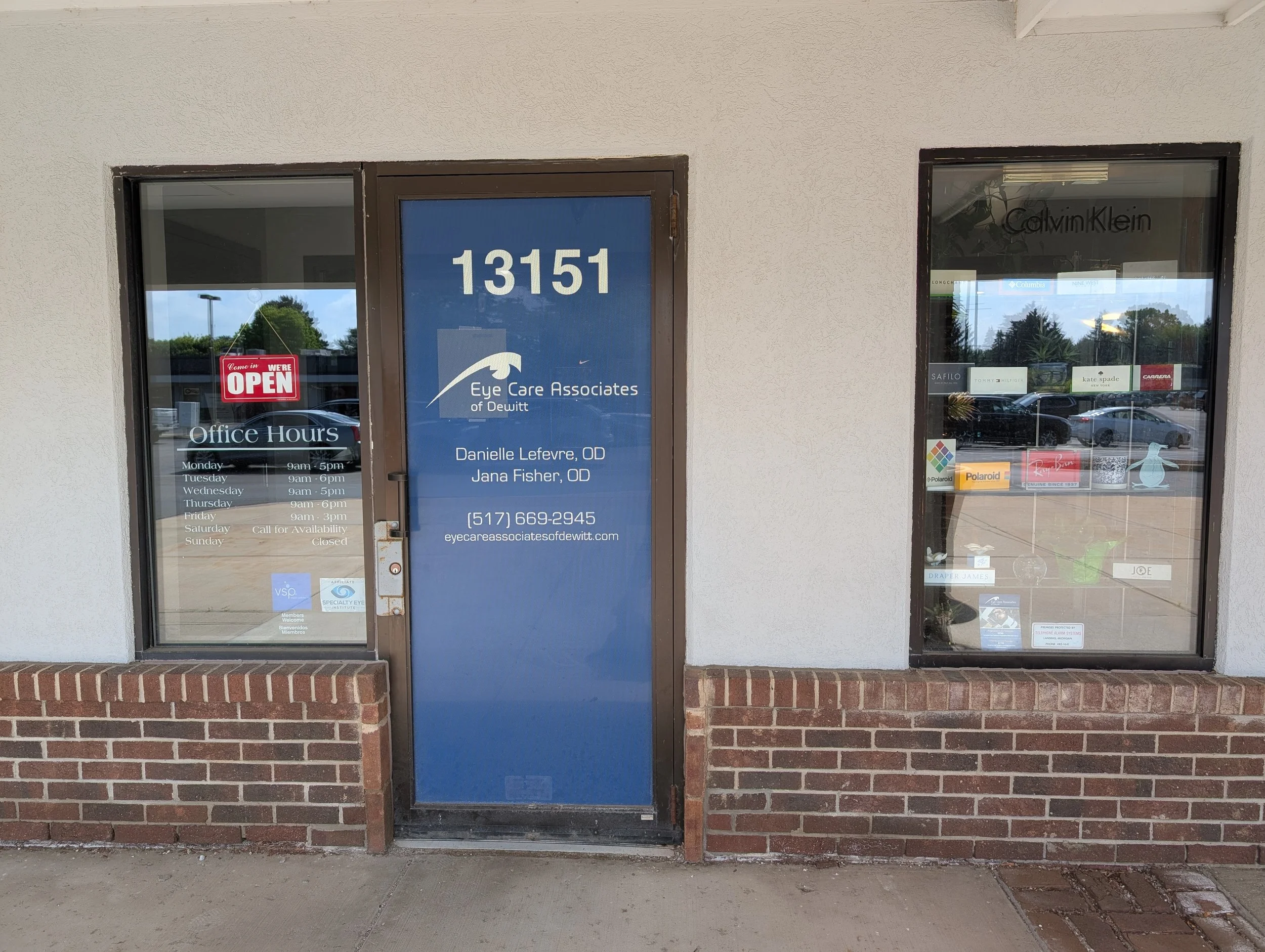 Storefront with two large windows and a door, one window has a red 'Come in We're OPEN' sign, and the door displays the business hours and contact information for Eye Care Associates of Dewitt.
