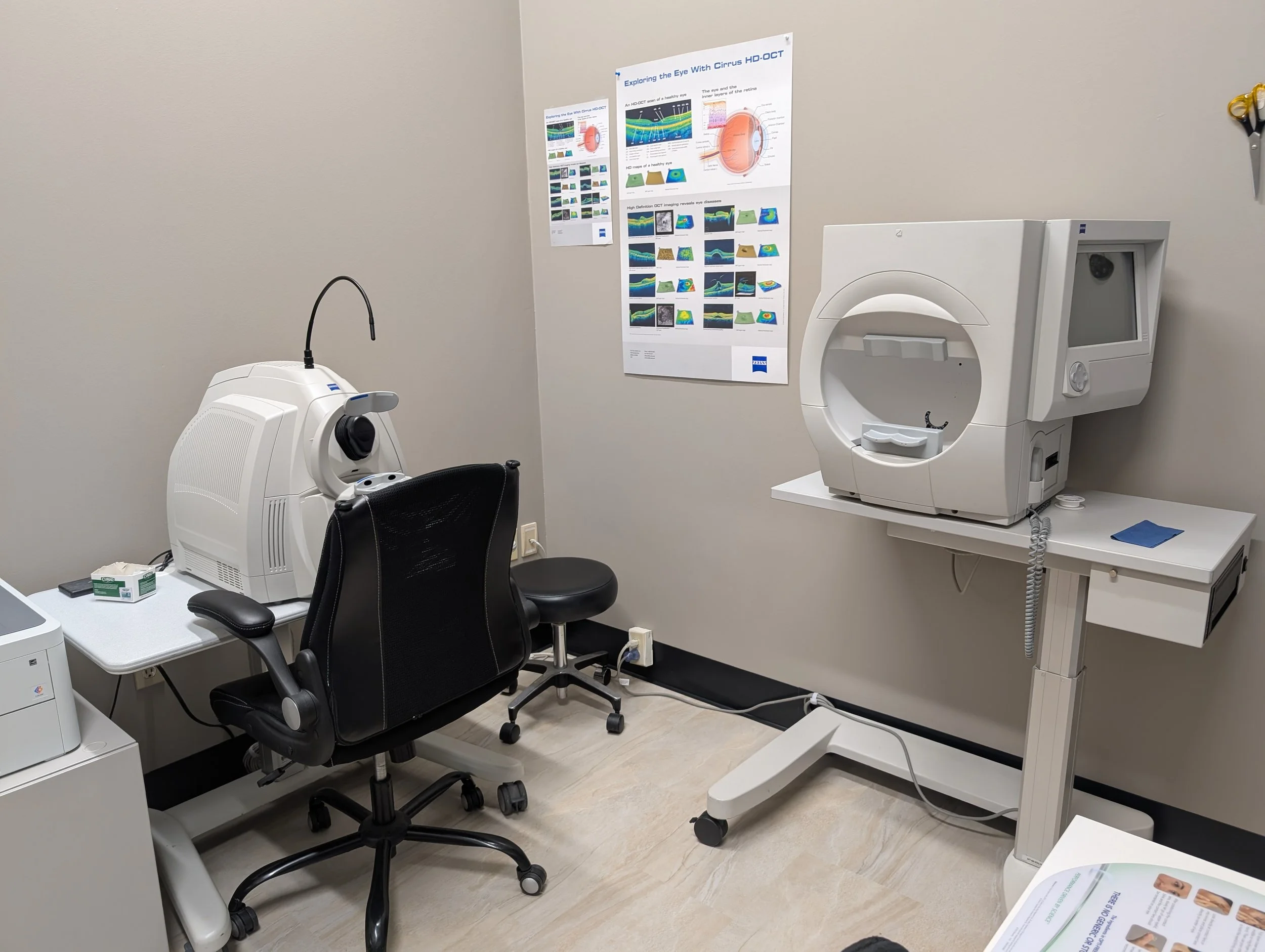 An ophthalmology examination room with a specialized eye imaging machine on the right, a black office chair, a small white table with an eye exam device and boxes on it, a black swivel chair, and informational posters related to eye health on the wal