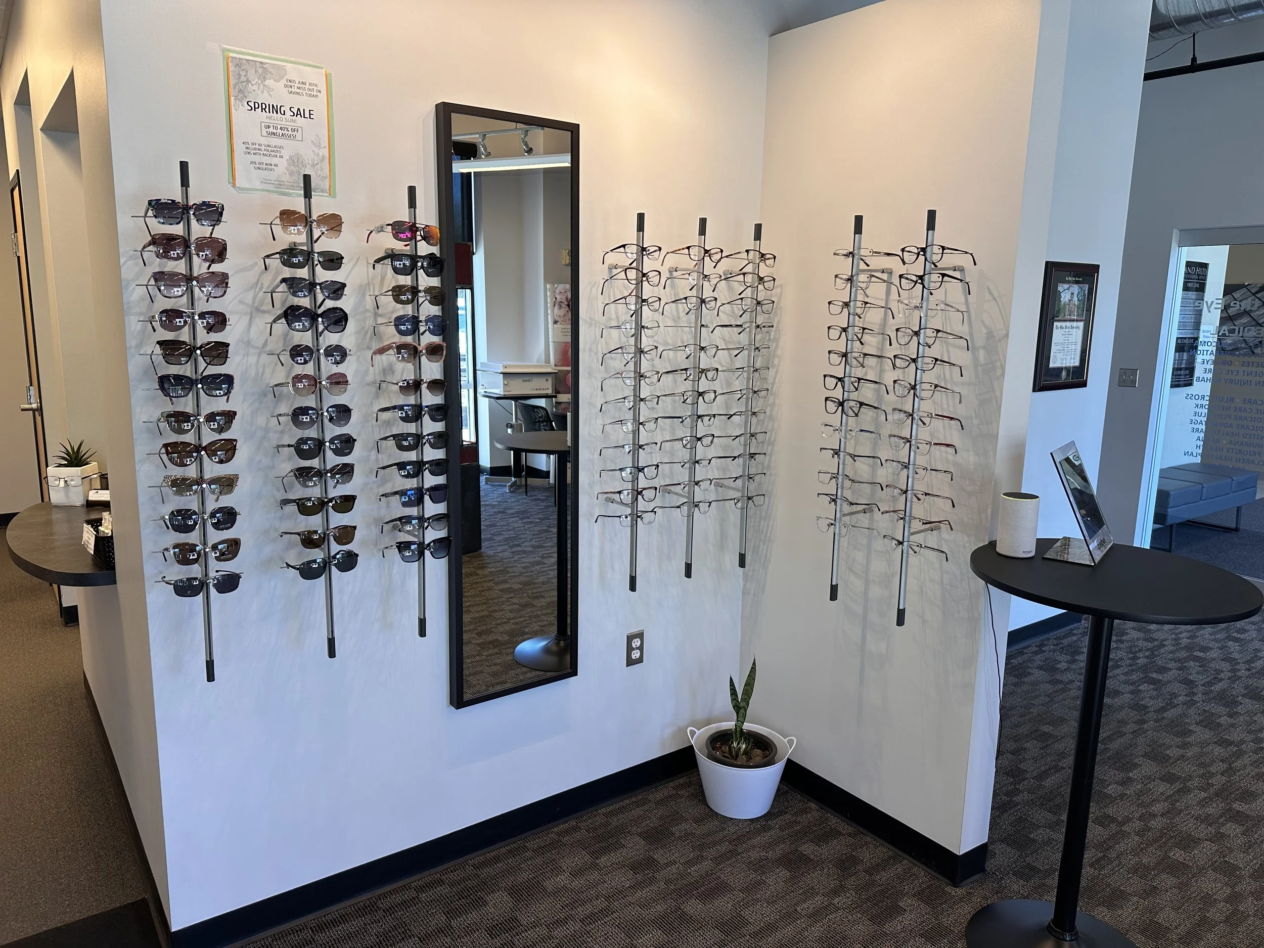 Inside an eyewear store, displays of sunglasses and eyeglasses on vertical racks, a full-length mirror, a small table with a plant, and promotional signs for spring sale and sunglasses discounts.