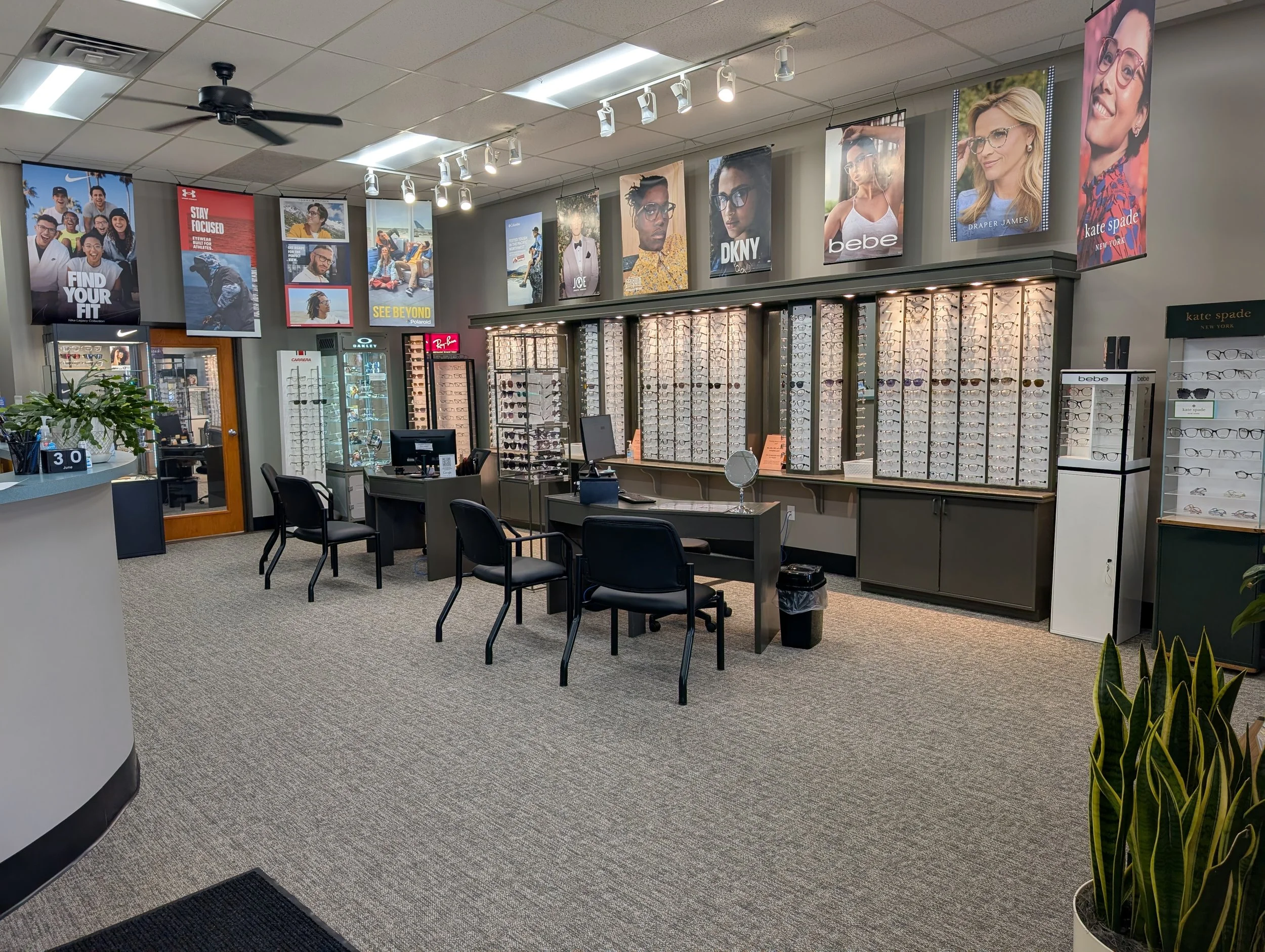 Inside an eyewear store with display cases filled with glasses, posters of models wearing glasses, a reception desk, chairs, and plants.