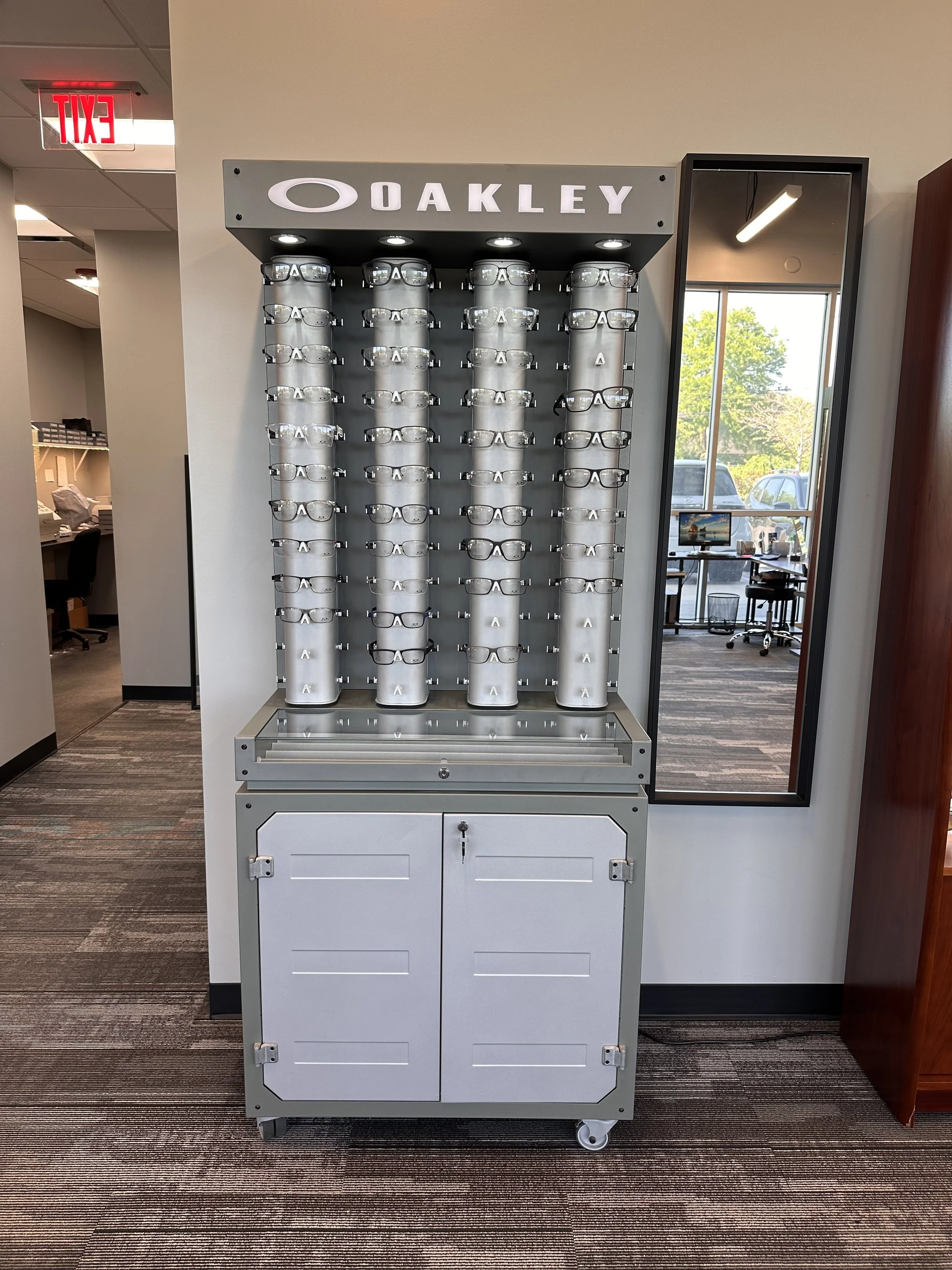 Display stand with multiple pairs of eyeglasses, branded with the Oakley logo, located in an office or retail environment.