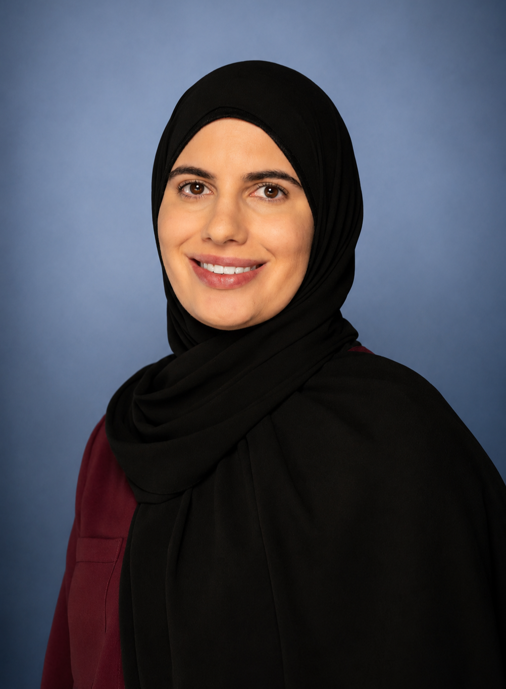 Portrait of a woman wearing a black hijab and maroon top, smiling against a blue background.