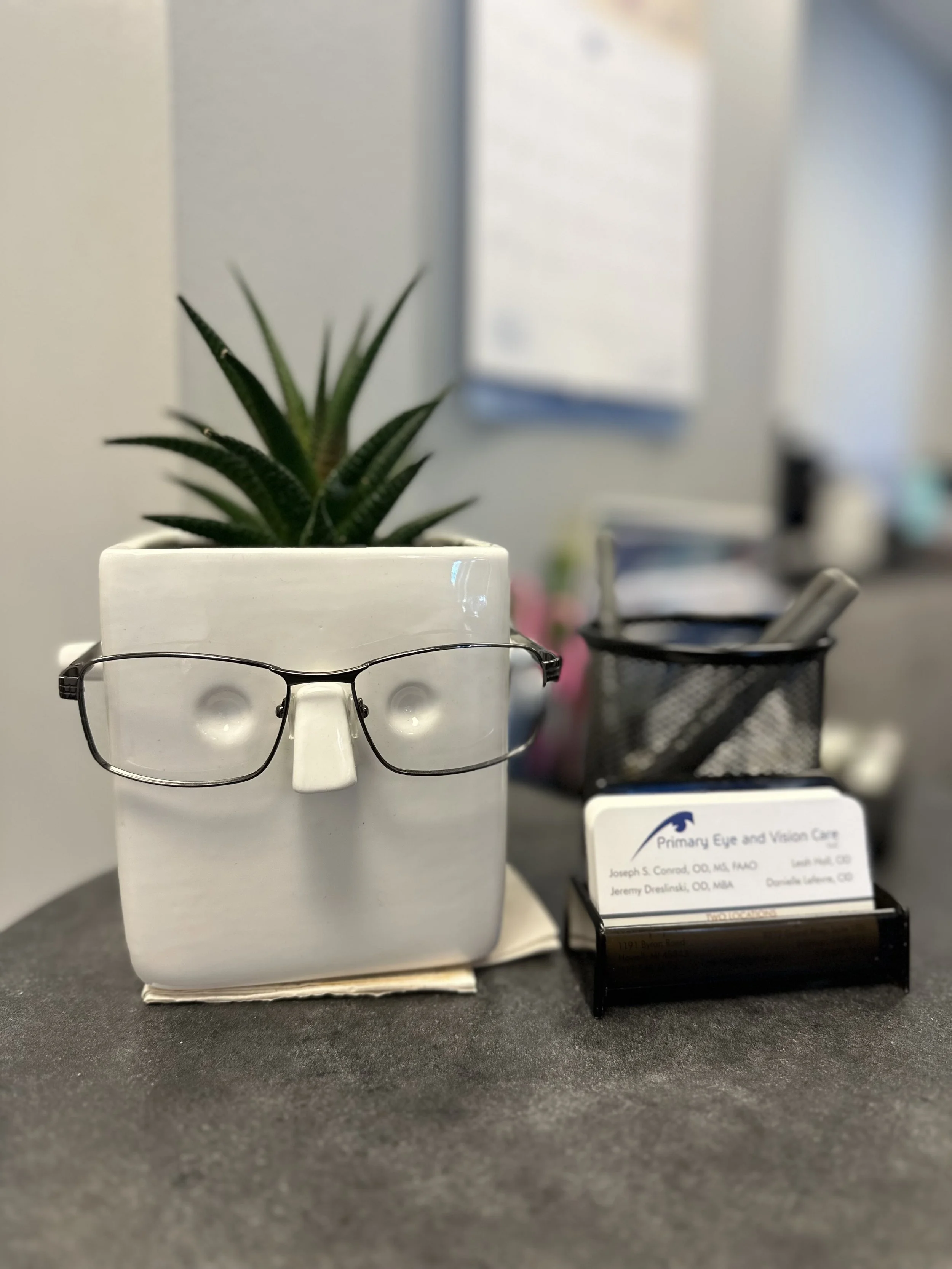 A small potted plant with green, spiky leaves sitting on a table. A pair of eyeglasses rest on the edge of the pot. In the background, there is a blurred office environment with a business card visible that reads "Primary Eye and Vision Care," and so