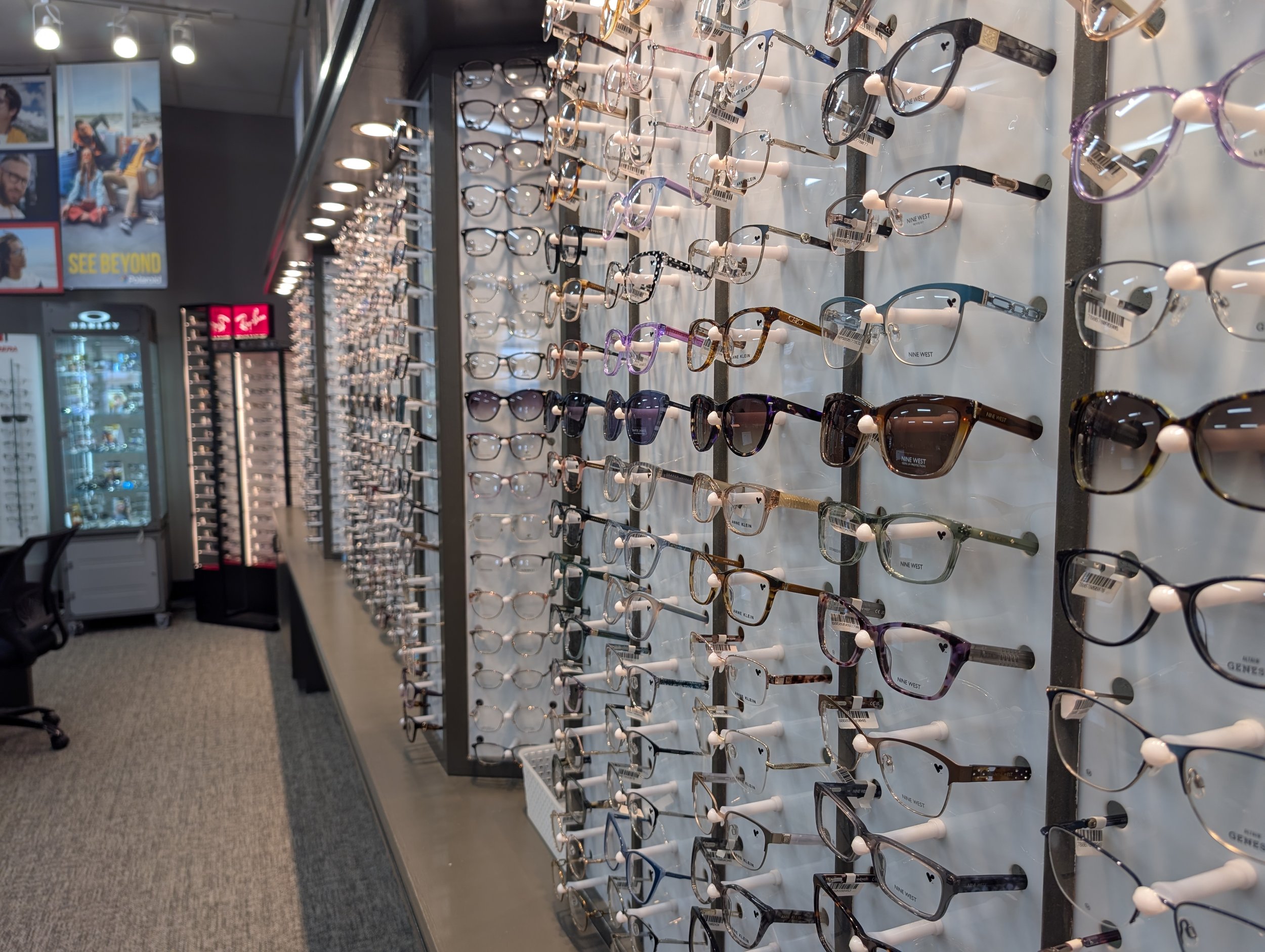Display of various eyeglasses and sunglasses on a retail store wall with a bright overhead lighting and posters in the background.