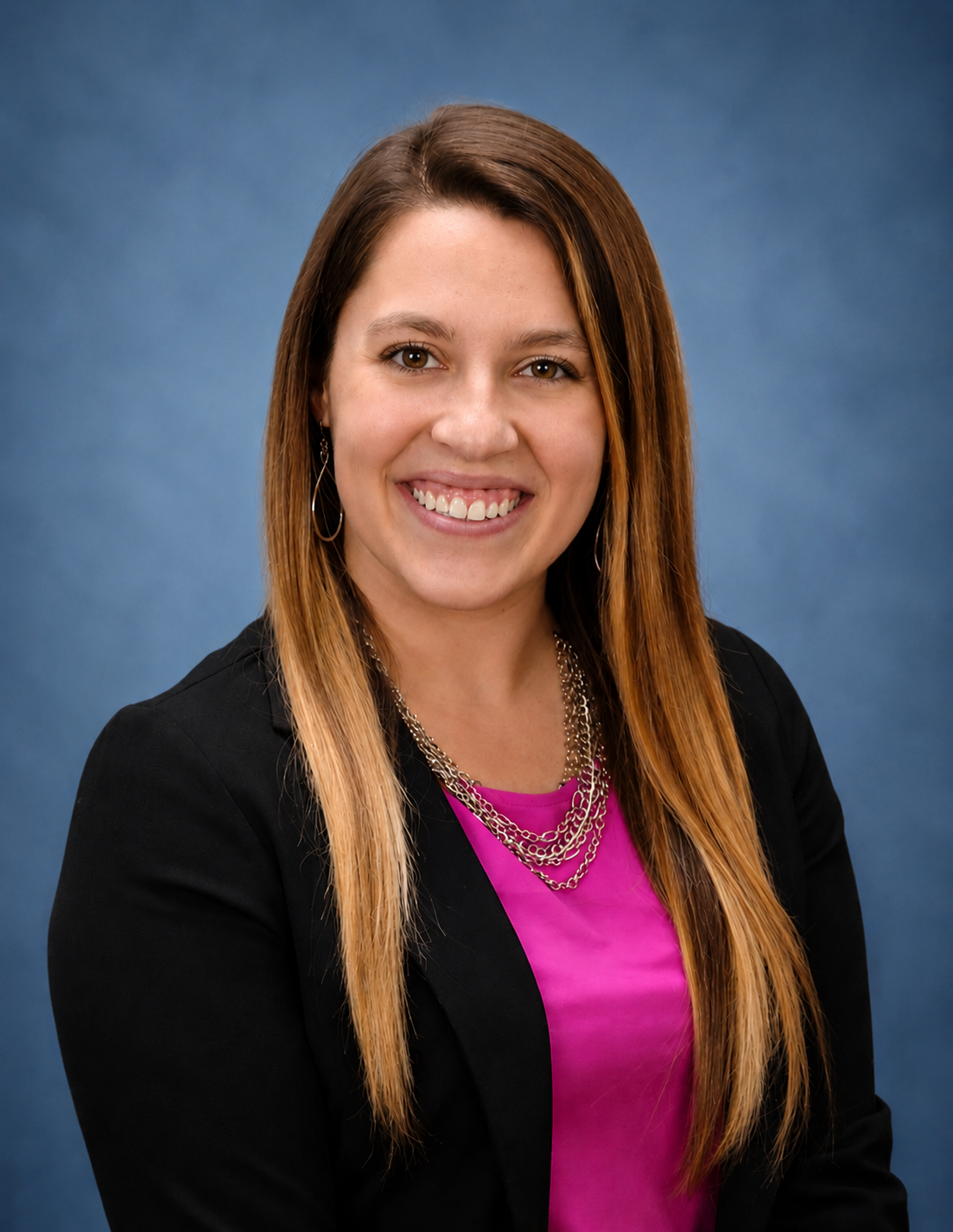 Professional headshot of a woman with long, light brown hair smiling, wearing a black blazer, pink top, silver necklace, and earrings against a blue background.