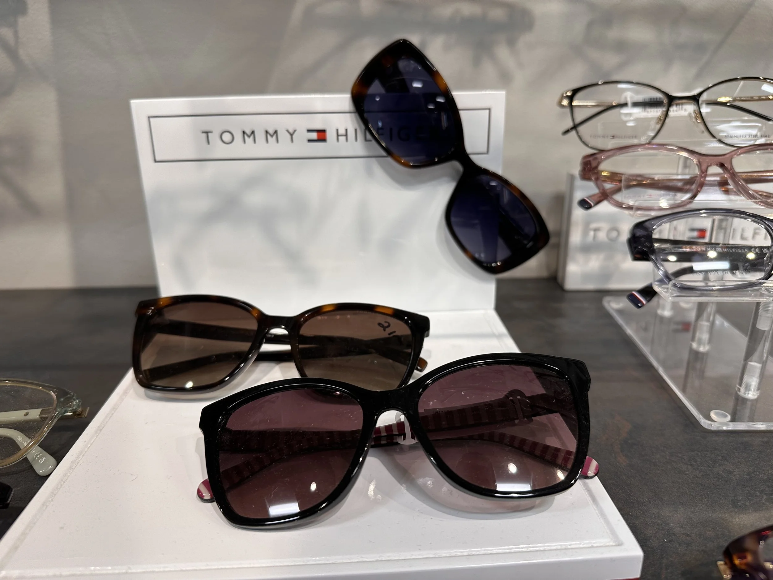 Display of sunglasses and eyeglasses on a retail shelf with a Tommy Hilfiger sign.